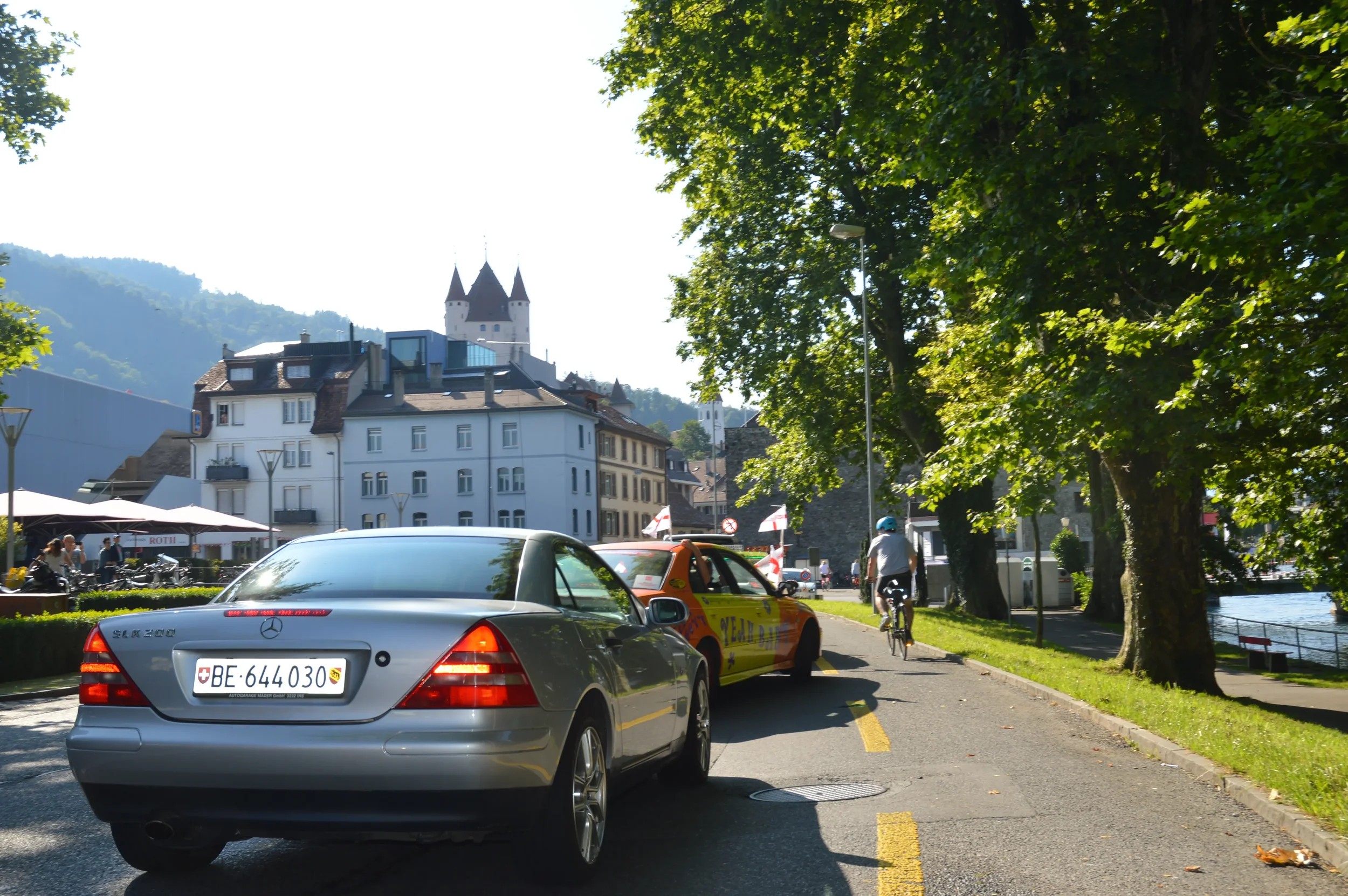 A street scene with a silver Mercedes-Benz car, a colorful race car, a cyclist, trees, and historic buildings with a castle on a hill in the background.