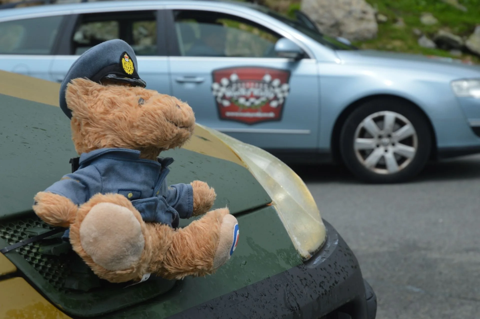 A teddy bear dressed as a police officer, wearing a police cap and uniform, sitting on the hood of a yellow vehicle. In the background, there is a silver car with a badge or logo on the door.