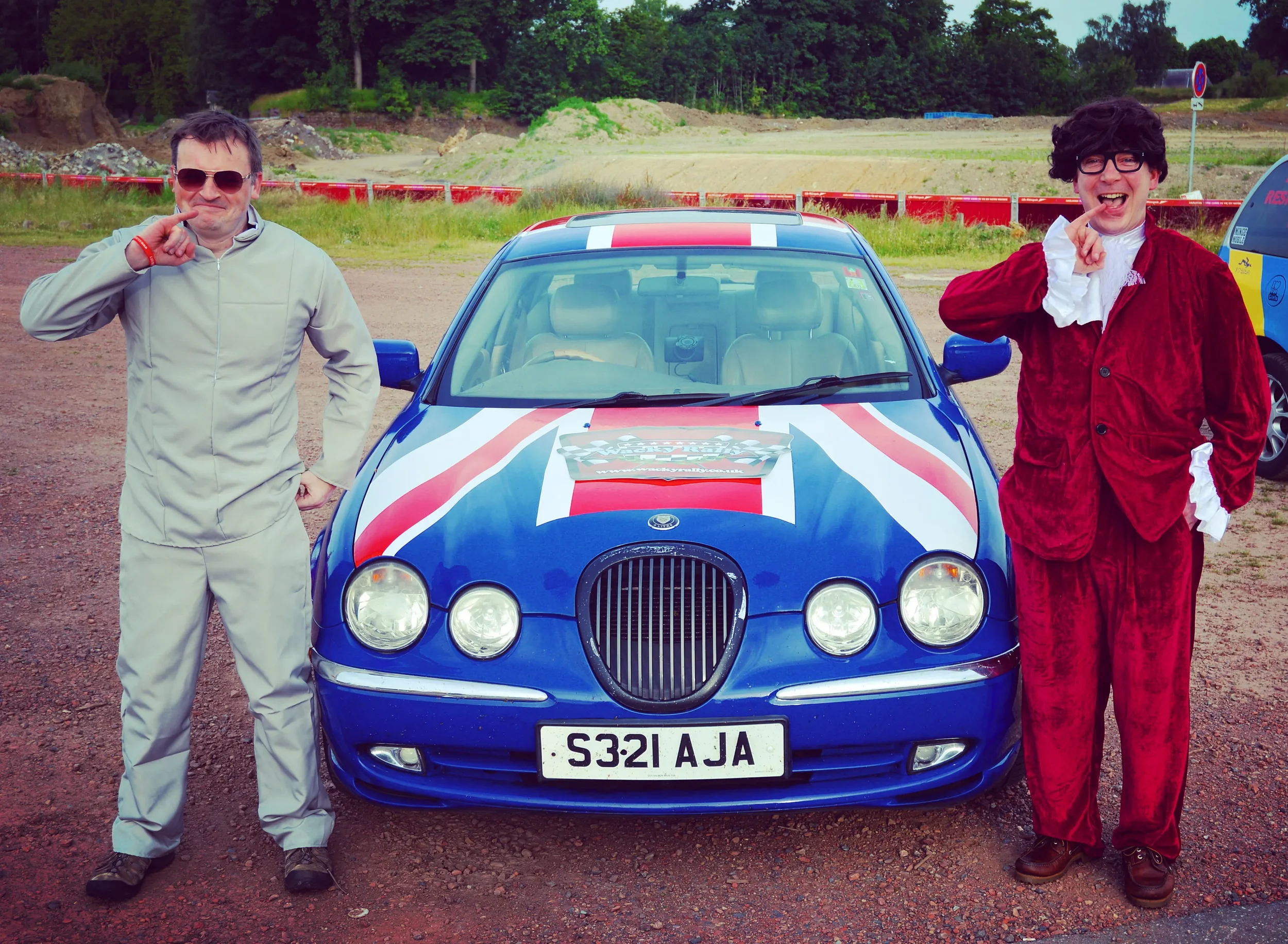 Two men standing next to a racing car painted with British flag colors at a race track, one in a beige racing suit and sunglasses, the other in a red velvet costume with curly black wig and glasses.