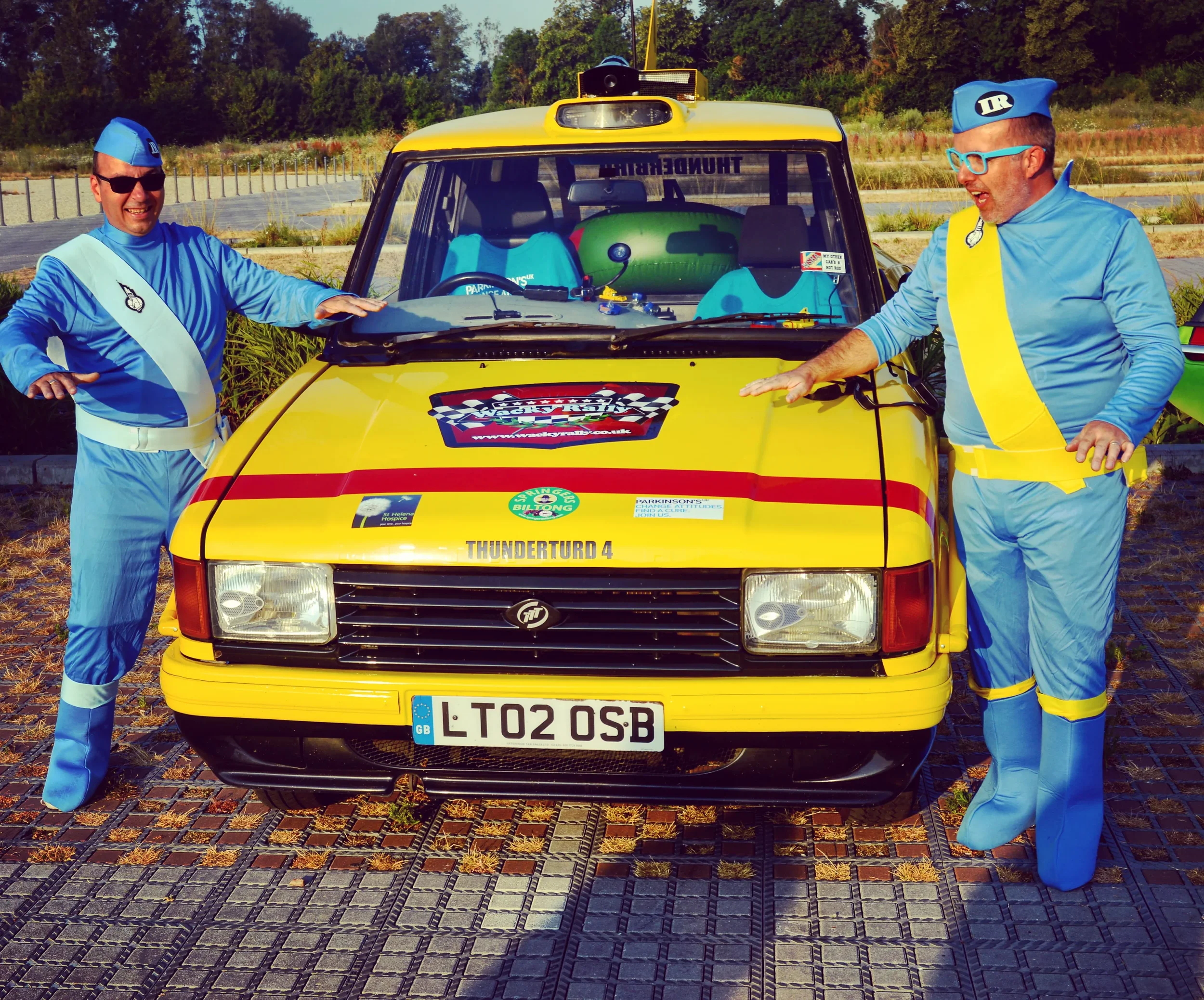 Two men dressed in blue astronaut costumes with yellow and white accents, standing on either side of a yellow car with racing stickers, in a parking lot with greenery in the background.