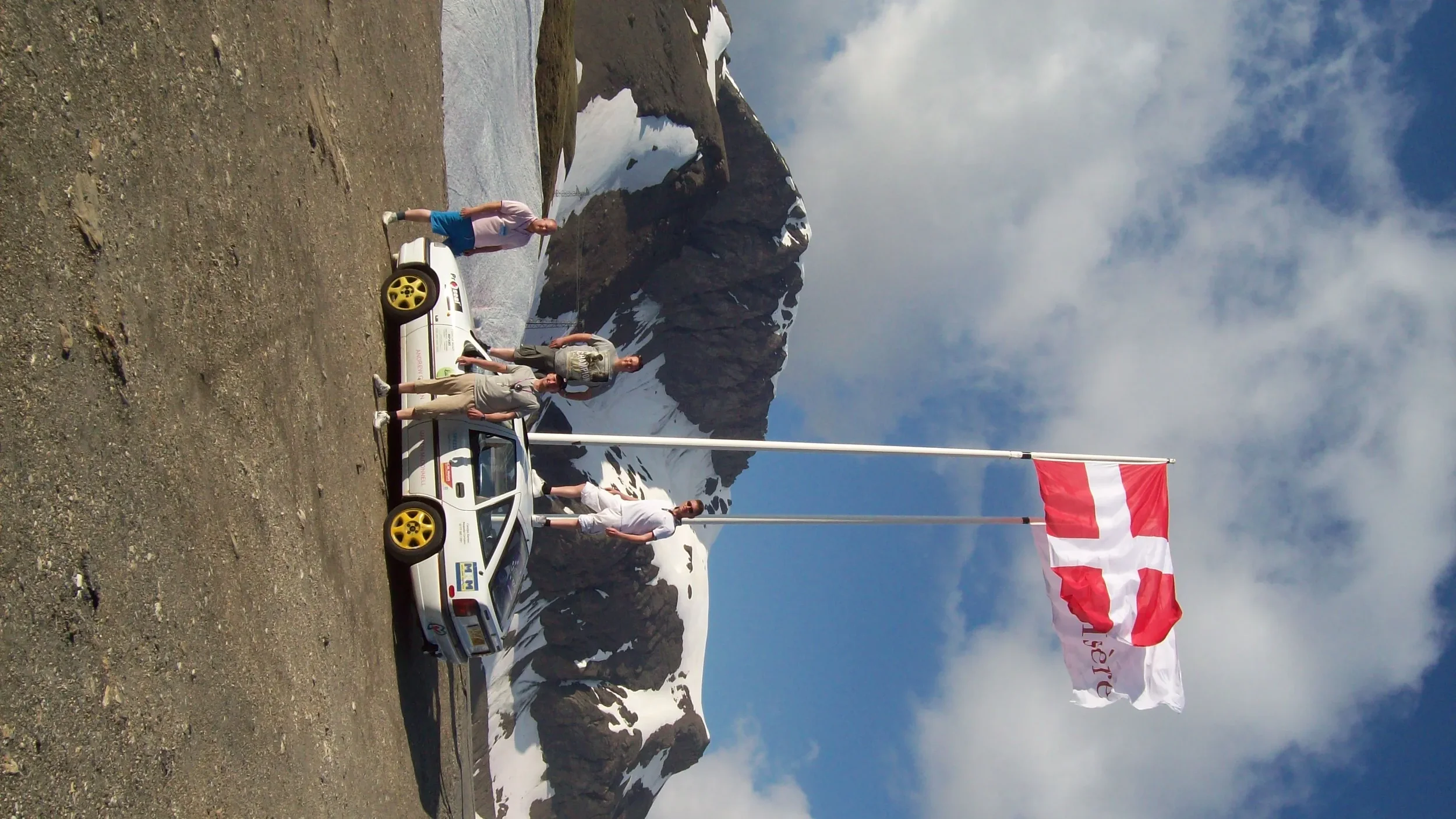 Four people standing next to two rally cars in a mountainous area with snow-covered peaks, holding a Danish flag.