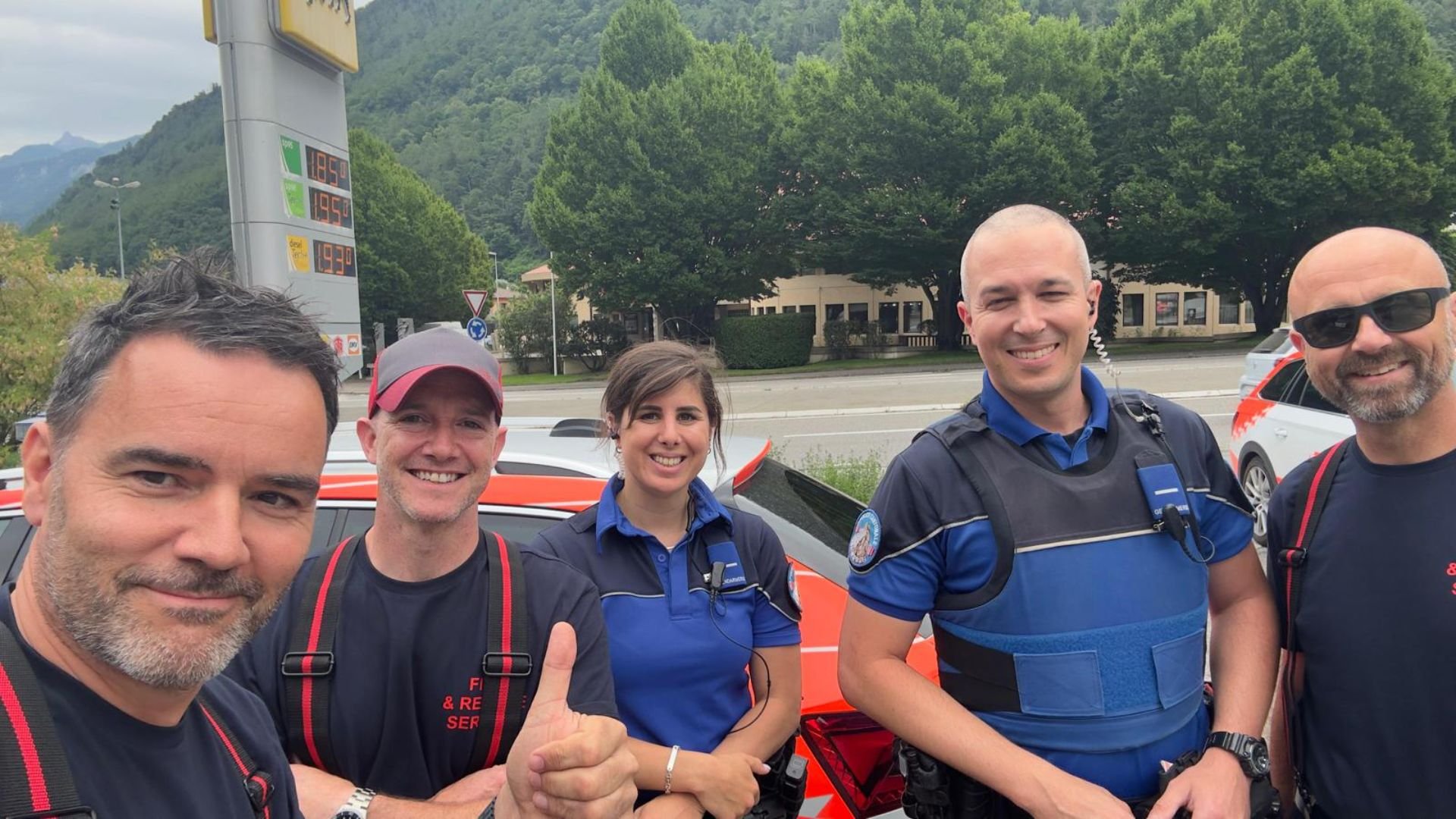 Five emergency responders, three men and two women, standing outdoors in front of an emergency vehicle, smiling at the camera. Behind them is a roadside with trees, a building, and vehicles, with a fuel price sign in the background.