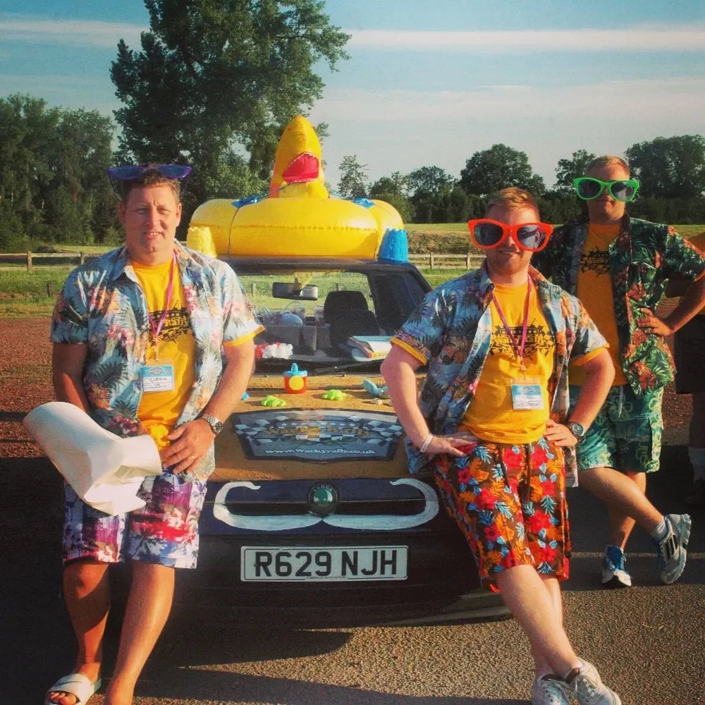 Three men dressed in colorful Hawaiian shirts and swim trunks, standing in front of a car with a large inflatable rubber duck on the roof, during daytime with trees in the background.