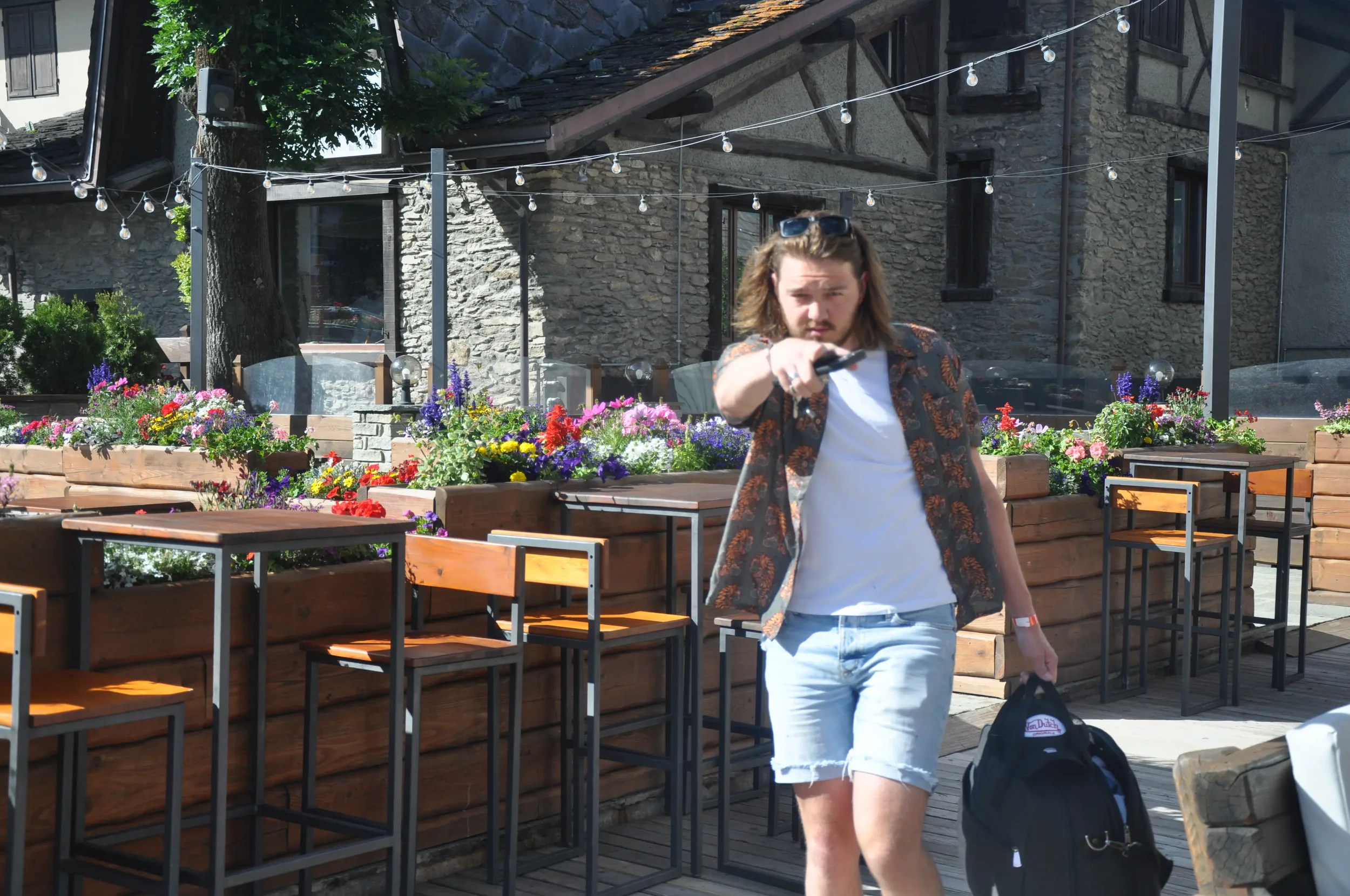 A man with long hair, wearing sunglasses, a patterned shirt, white T-shirt, and denim shorts, walking past an outdoor patio with flowers and tables, holding a black bag in his left hand and pointing a remote or object towards the camera.