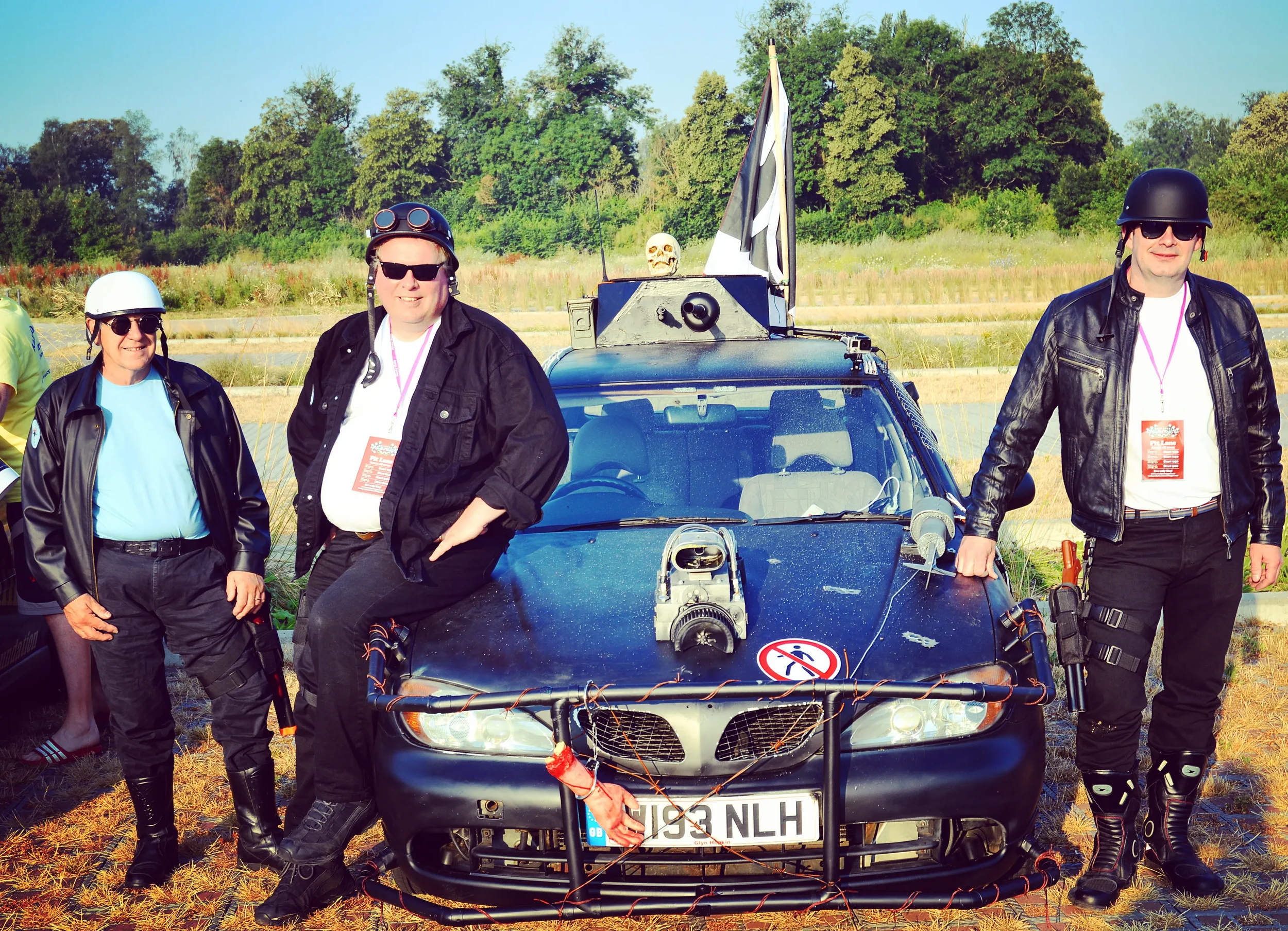Three people dressed as police officers standing in front of a black police car with a screen on top and a 'no walking' sign on the front bumper, outdoors on a sunny day with trees in the background.