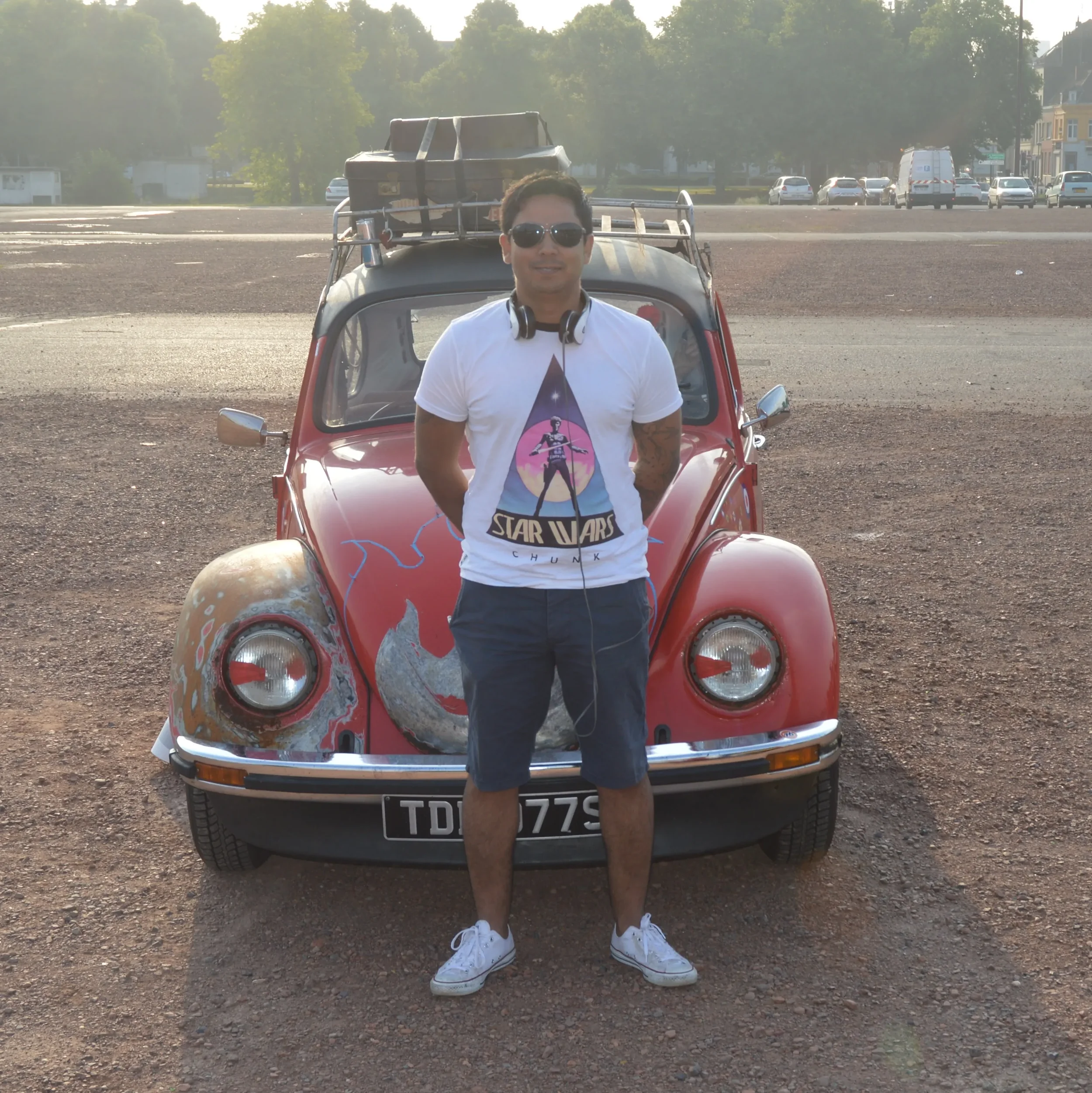 A man wearing sunglasses, a white Star Wars T-shirt, and shorts standing in front of a vintage red Volkswagen Beetle with artwork of a dragon on the front, in an open parking lot during daytime.