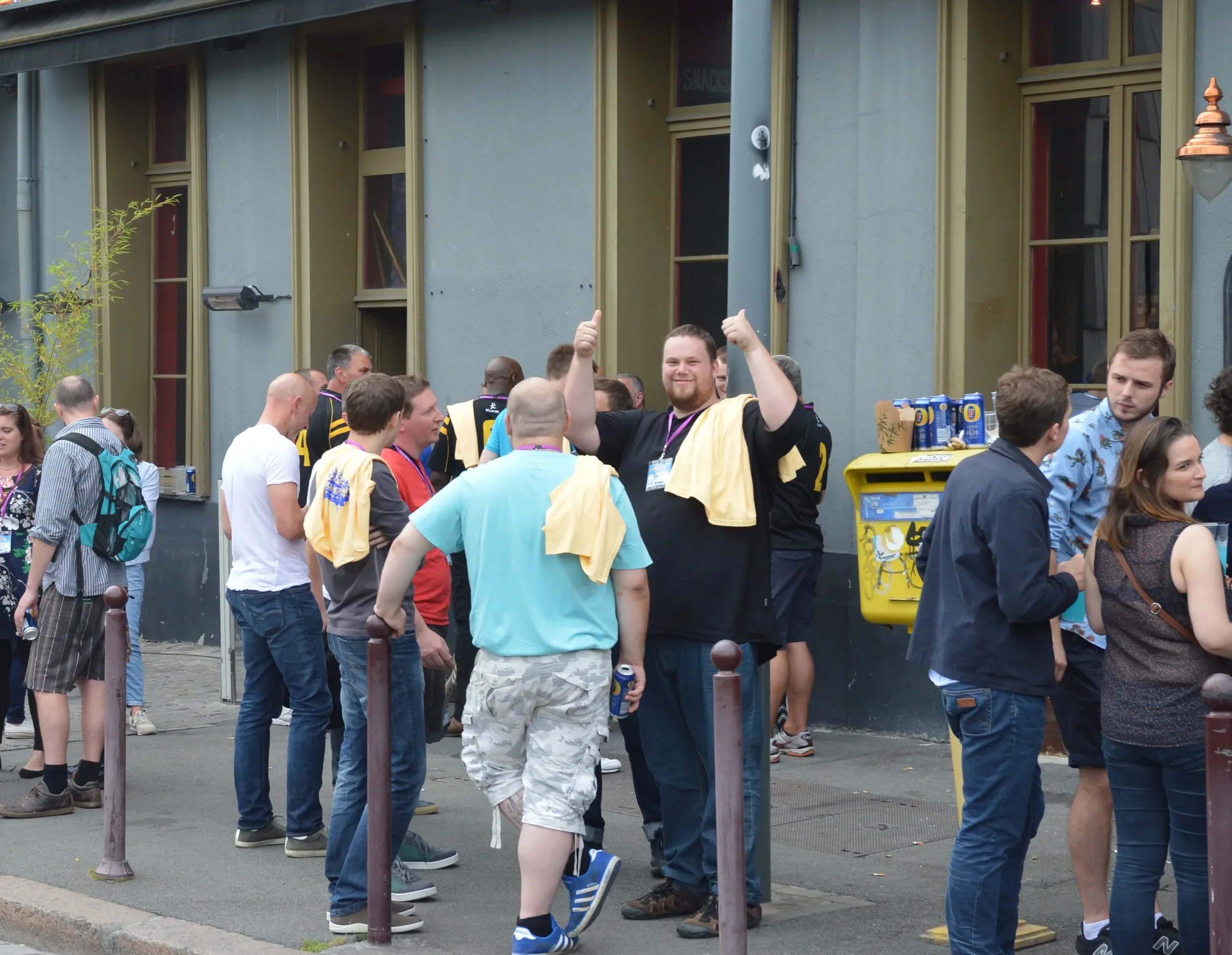A group of people gathered outdoors in line against a building, with one man in a black and yellow uniform smiling and raising his thumbs up.
