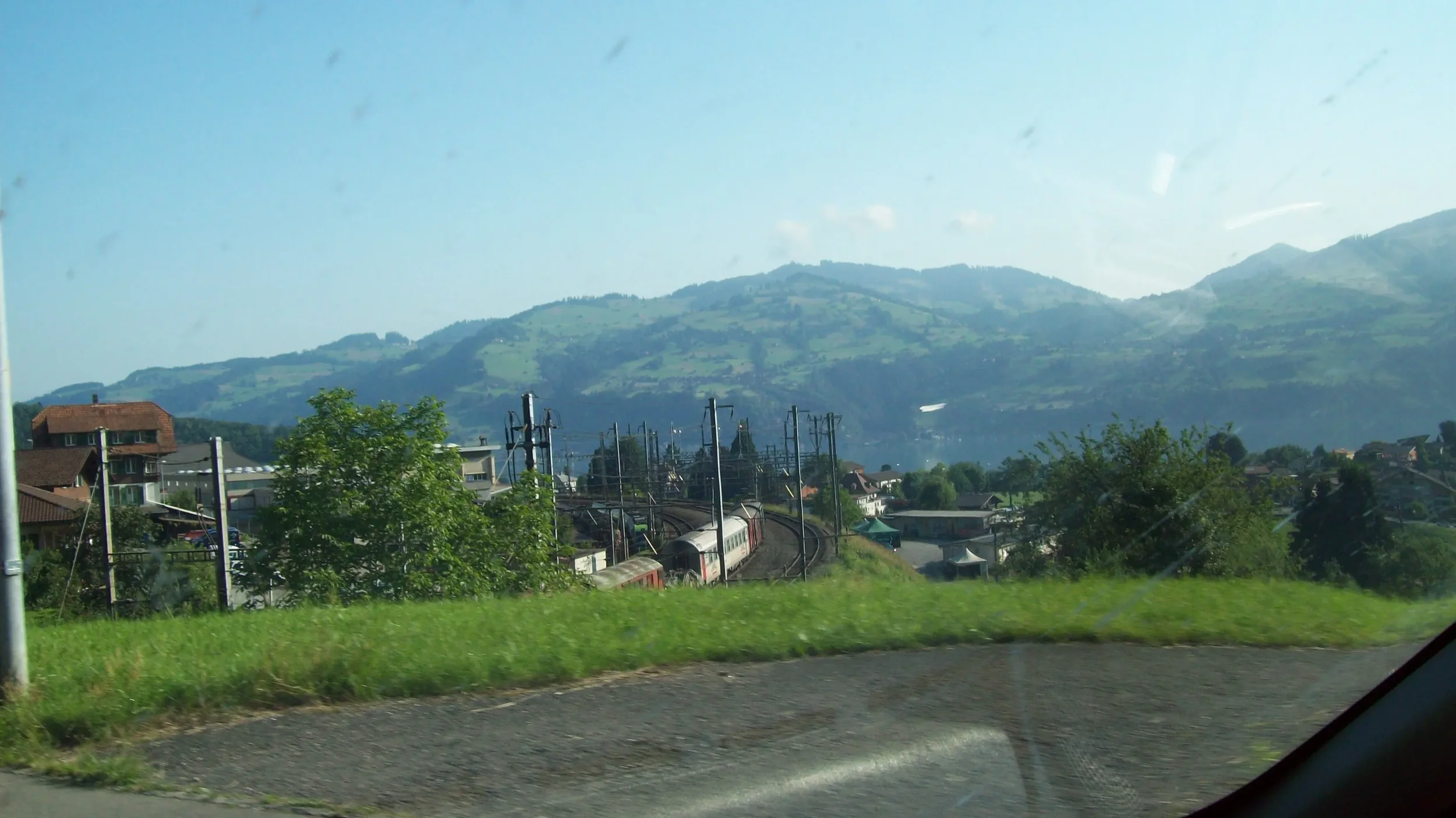 Train traveling through a scenic landscape with green hills and houses in the background, viewed from a moving vehicle.