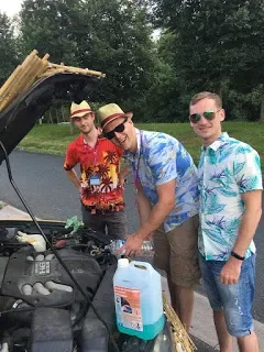 Three young men standing beside an open car hood, working on the engine in a park with trees and grass in the background, all wearing summer shirts and sunglasses.