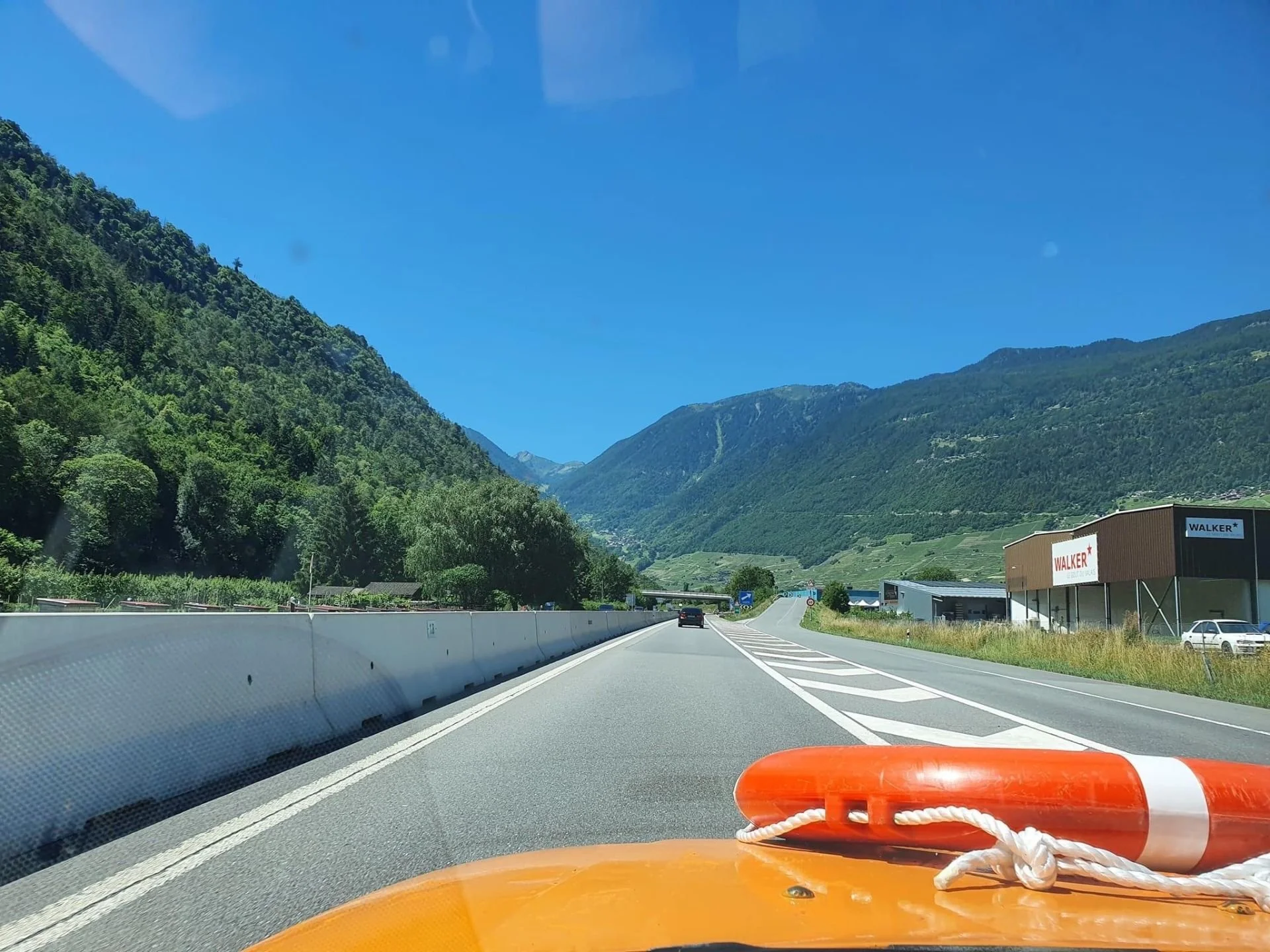 View of a highway surrounded by green mountains on a clear, sunny day, with orange safety equipment on the vehicle's cabin roof and a storefront on the right side of the road.