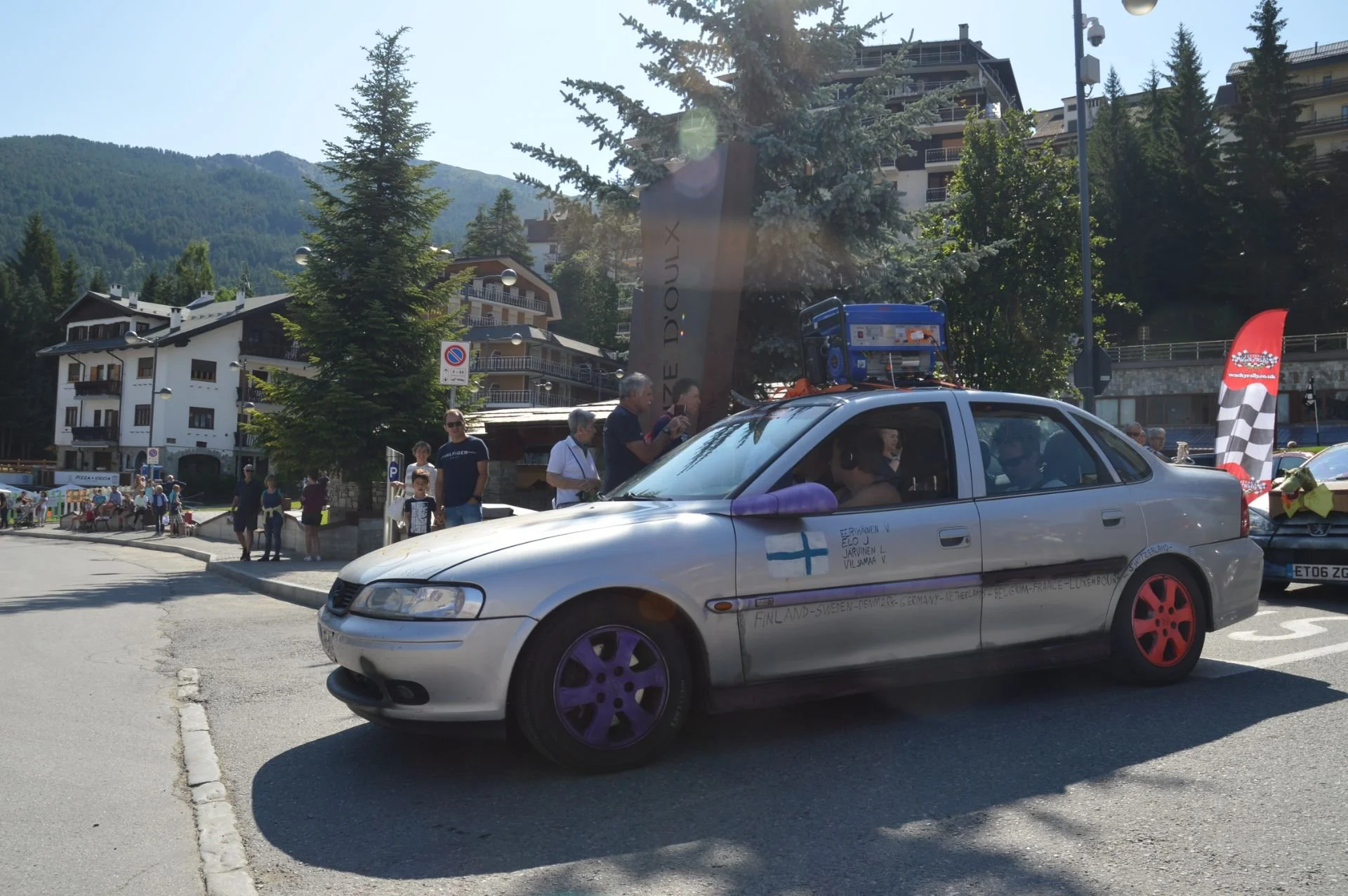 A silver car with purple and red wheels parked in a mountain town square, with people standing and sitting nearby, surrounded by trees and buildings, with hills in the background.