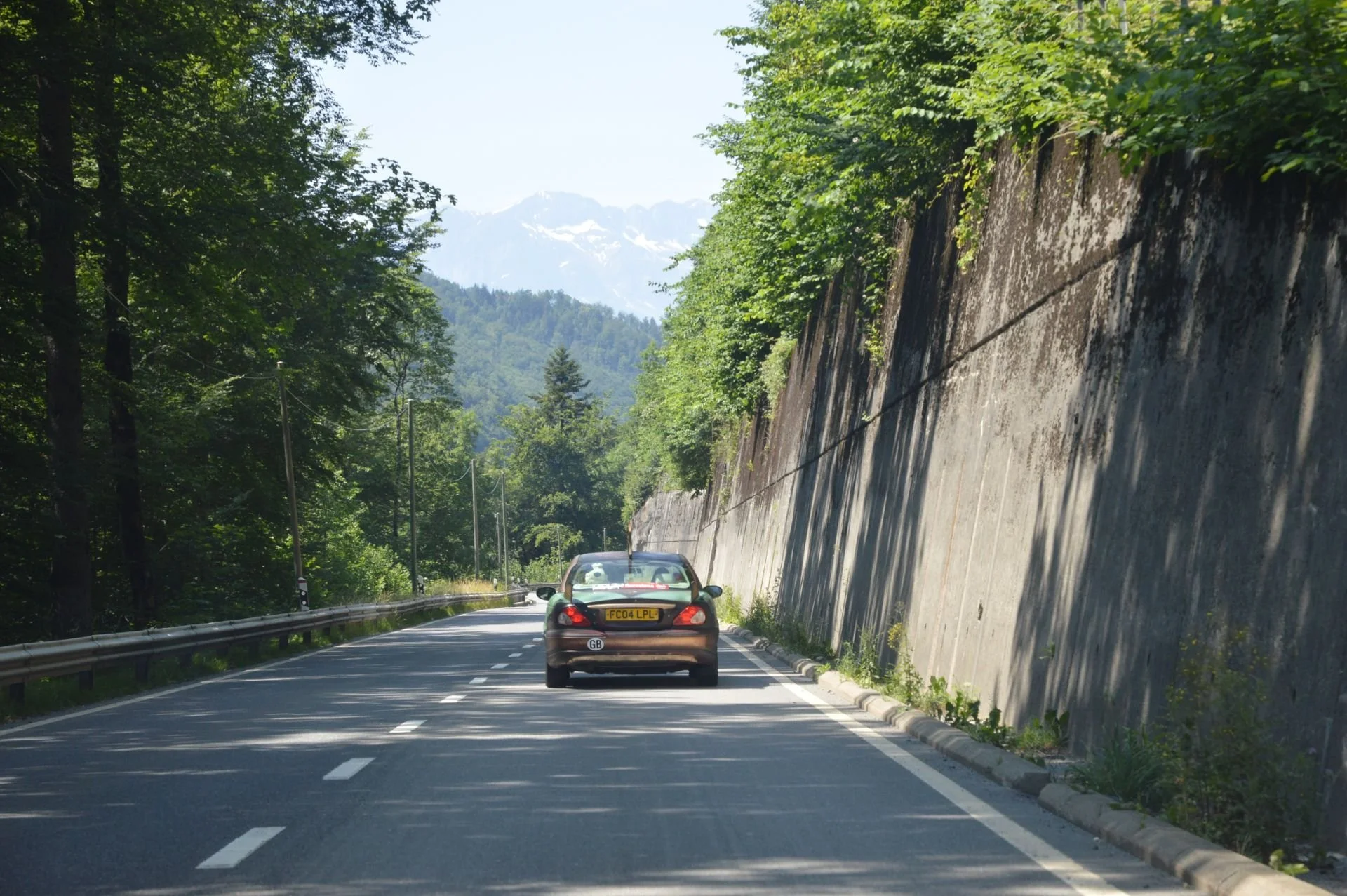 A black car driving on a mountain road with a concrete wall on the right and trees on the left, with mountains visible in the background.