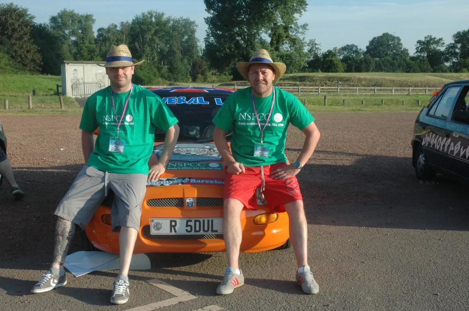 Two men wearing green NSPCC t-shirts and straw hats, seated on the front of a bright orange rally car with a black hood, parked in an outdoor area with trees and a wooden fence in the background.