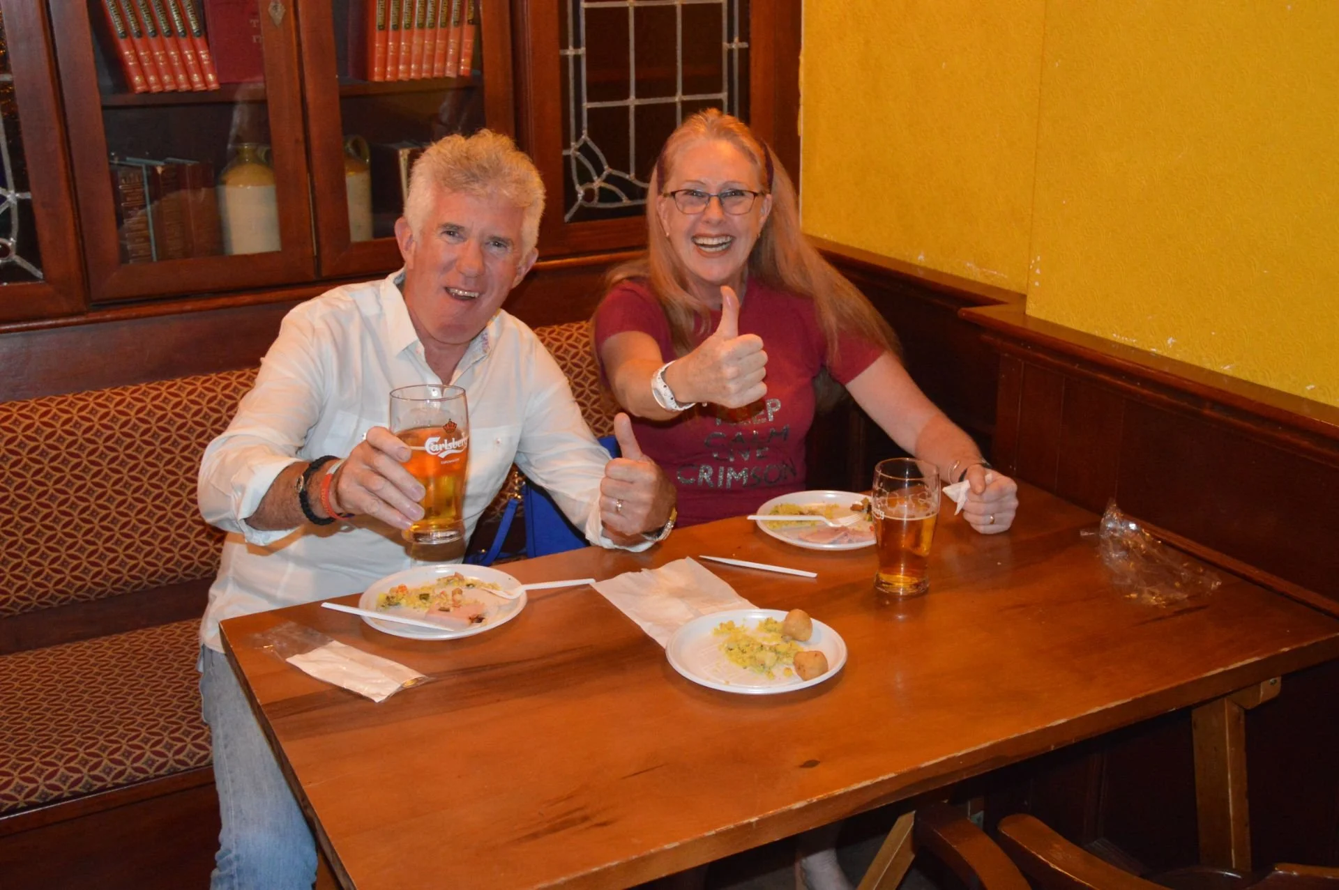 A man and woman sitting at a wooden table in a restaurant, smiling and giving thumbs-up gestures. They have plates with food and glasses of beer. The man has short gray hair and is wearing a white shirt, while the woman has long red hair, glasses, an