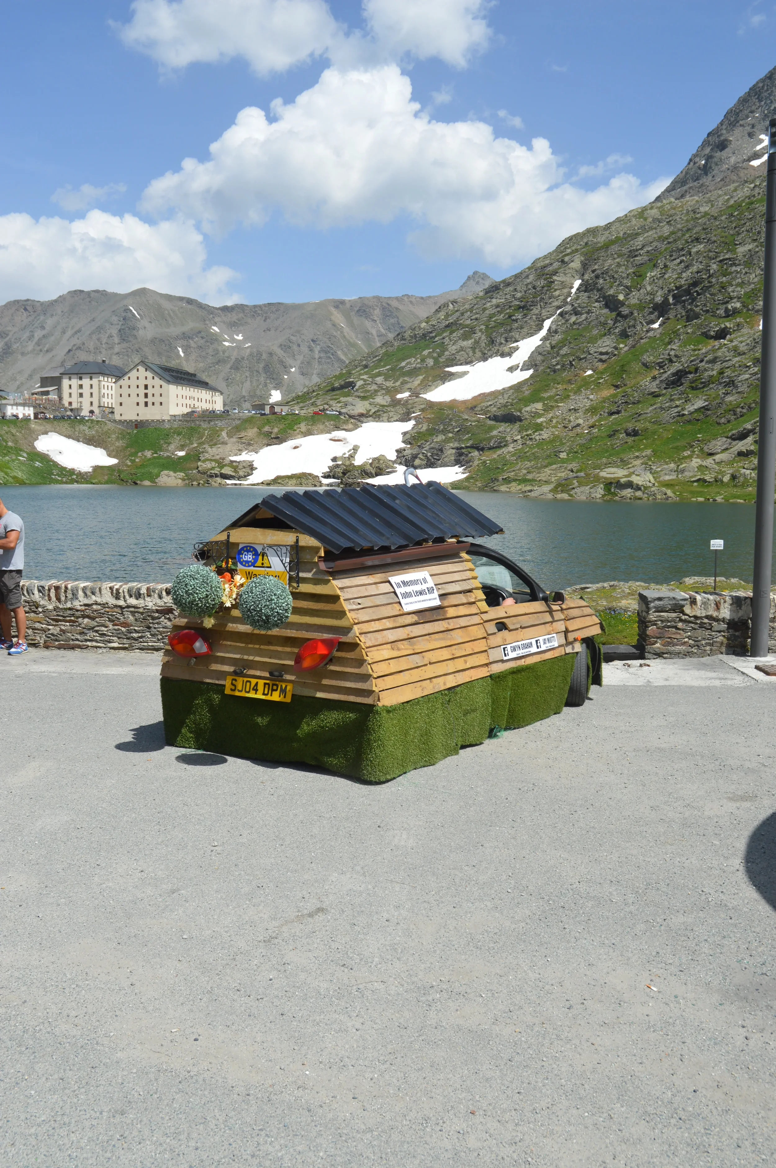 A small car decorated with grass and wooden slats resembling a tiny house, parked by a mountain lake with snow patches on the hills, and a building in the background.