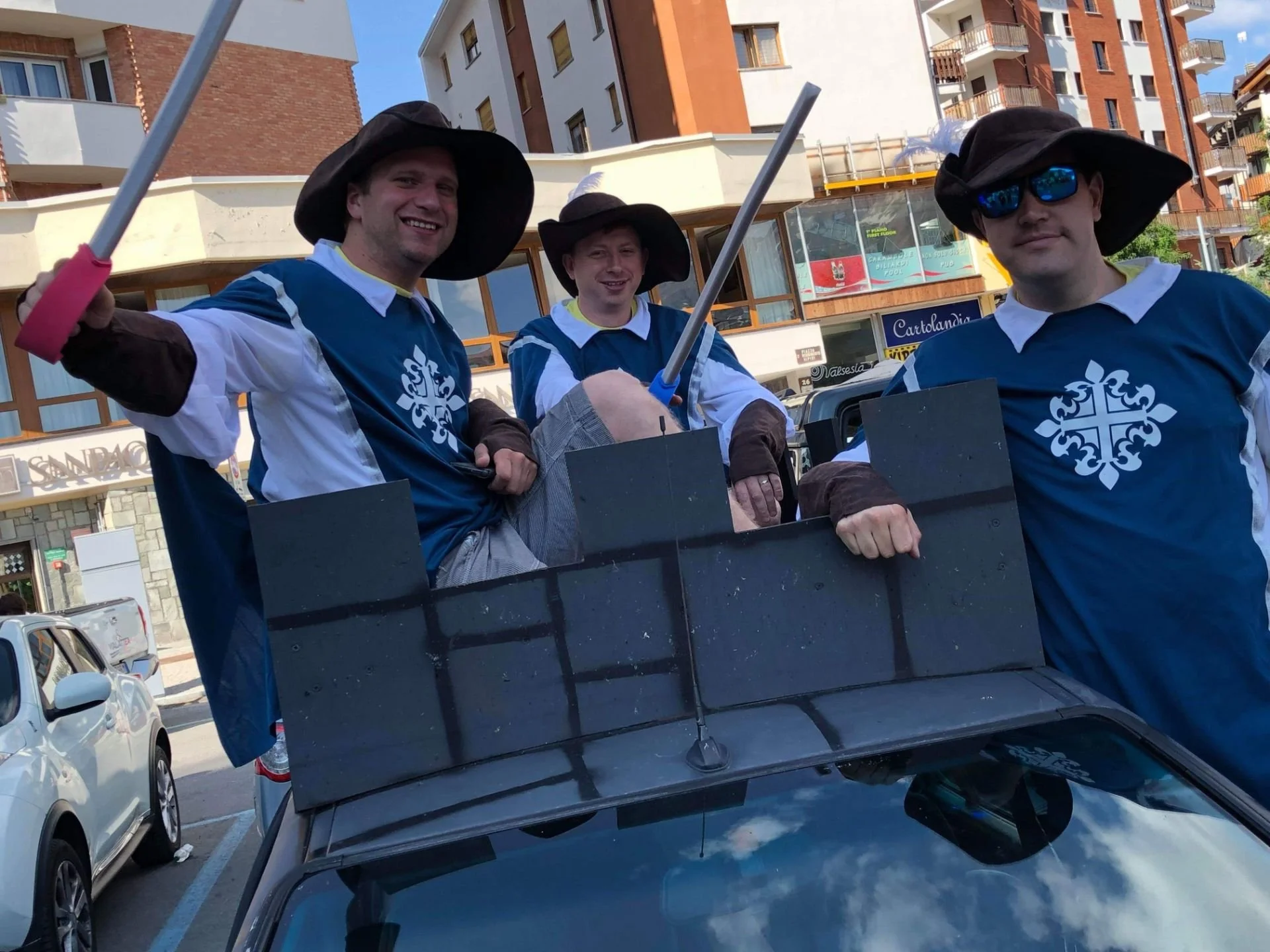 Three men dressed as medieval knights with blue and white tabards, black wide-brimmed hats, and sunglasses, riding on a float with a small black fortress replica during a parade in an urban area with buildings and parked cars.