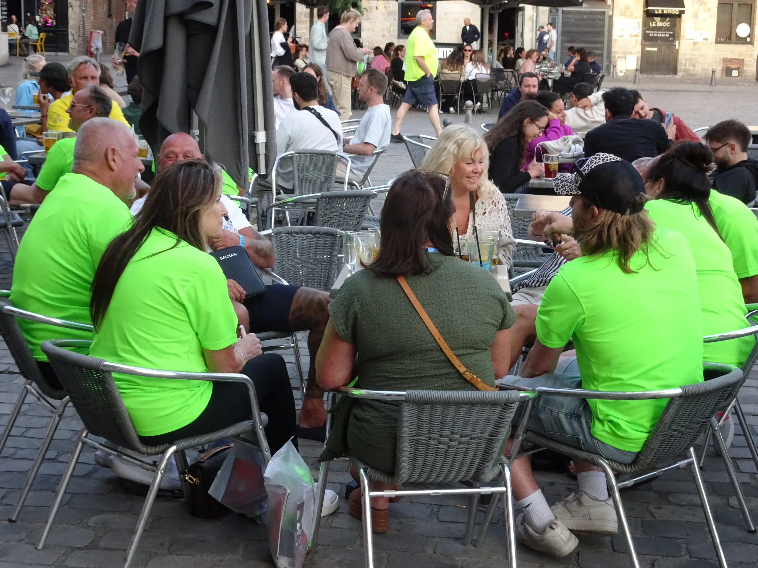 A group of people seated outdoors at a restaurant, some wearing neon green shirts, engaging in conversation, with additional people in the background at tables.