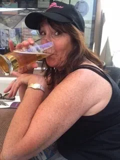 Woman wearing a black cap and sleeveless top, holding a beer glass near her lips, sitting outdoors.