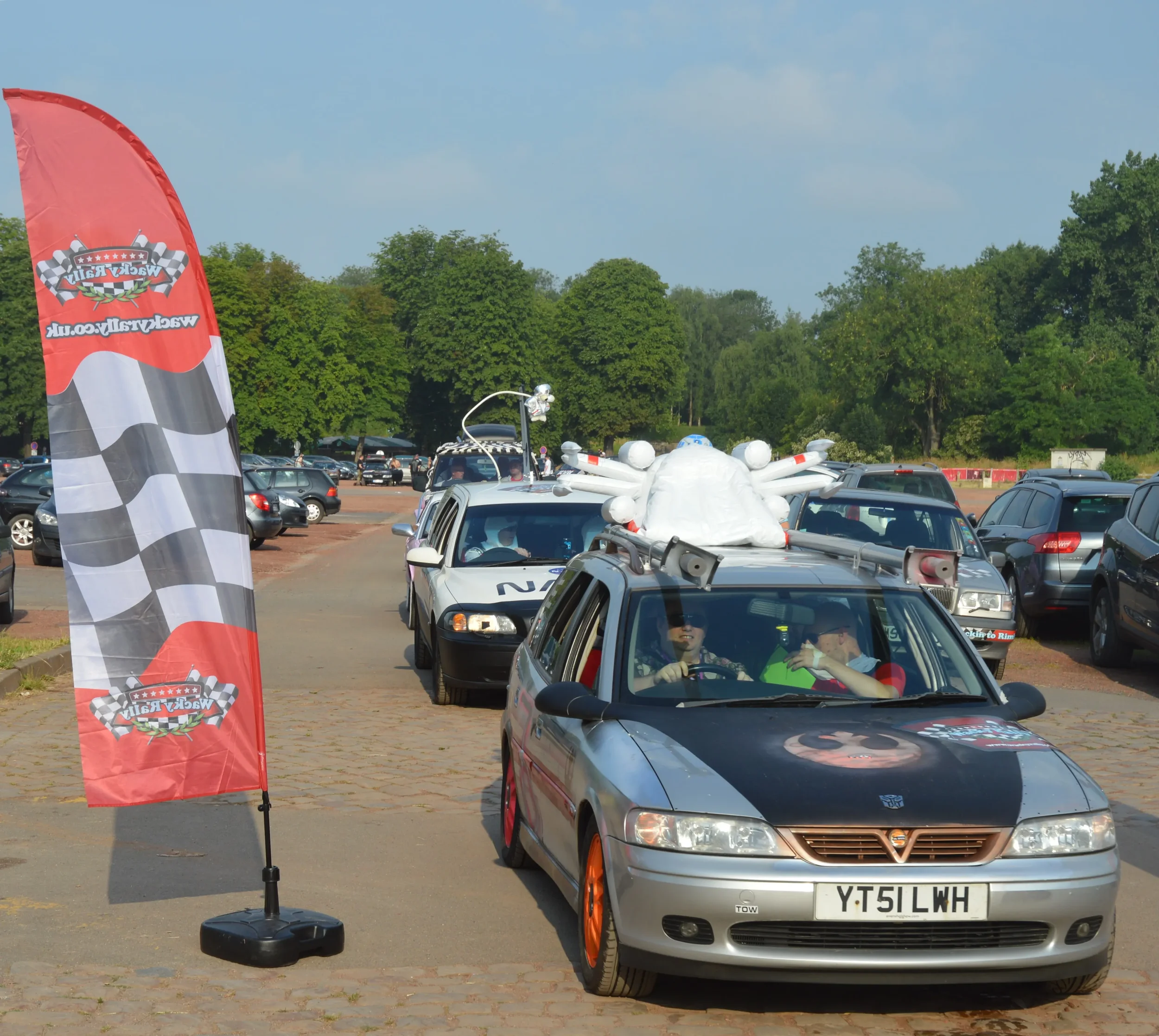 Line of cars decorated with racing-themed decorations at a race event, with a red flag displaying checkered flags and a race logo, and trees in the background.