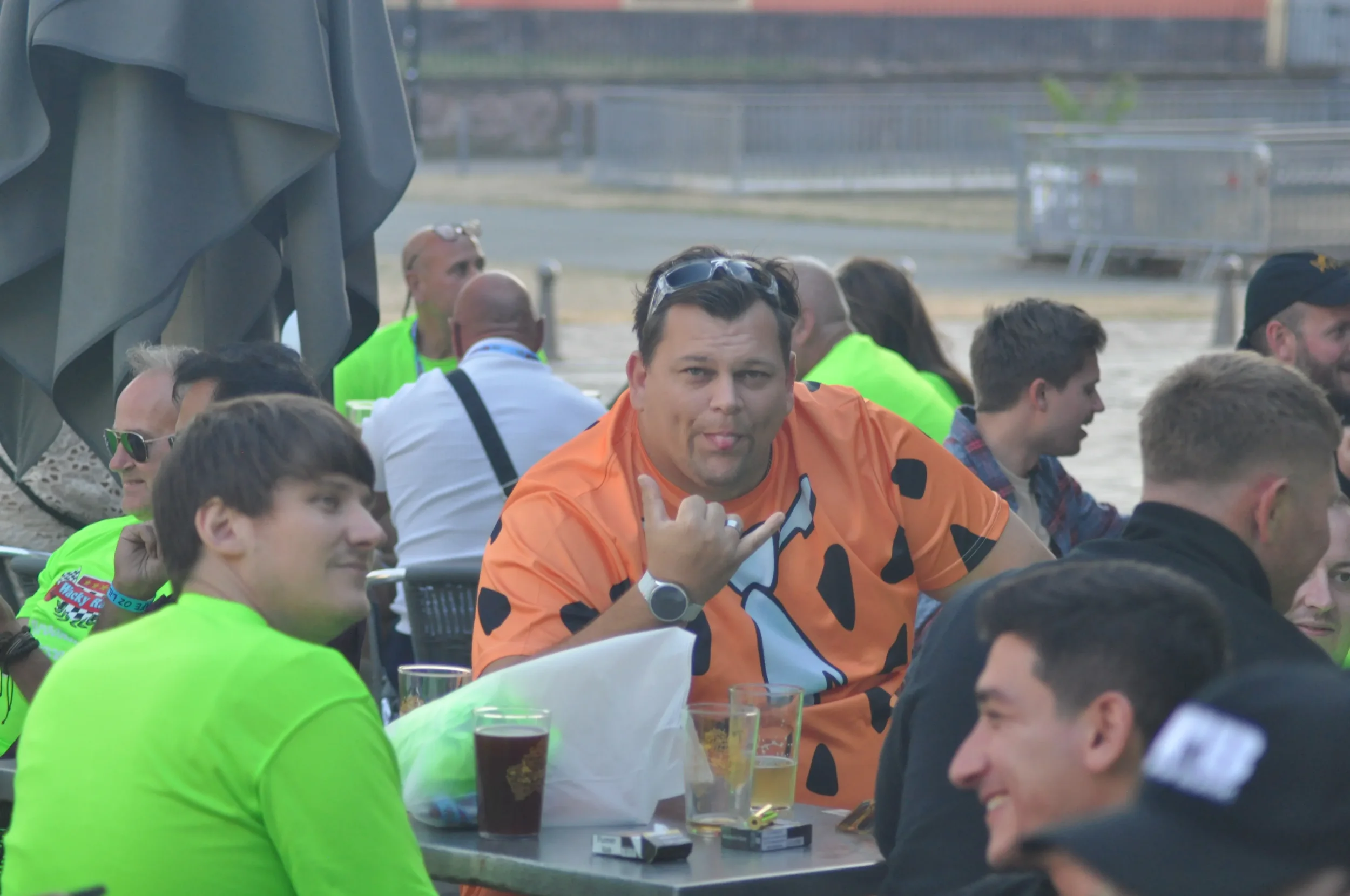 Group of people sitting at outdoor tables, some smiling and talking, some with drinks, one man making a shaka sign, wearing an orange shirt with a cow print, sunglasses on his head, in a casual gathering.