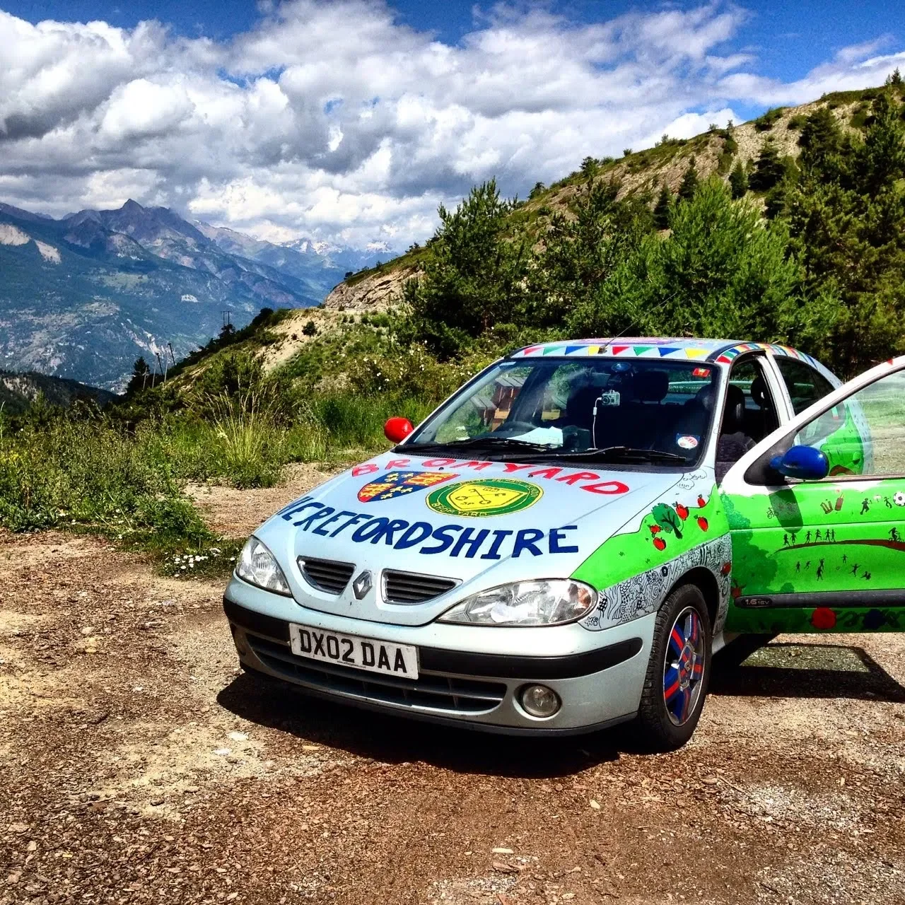 A decorated silver Renault car with colorful graphics and text, parked on a dirt road in a mountainous area with green hills, trees, and cloudy sky in the background.