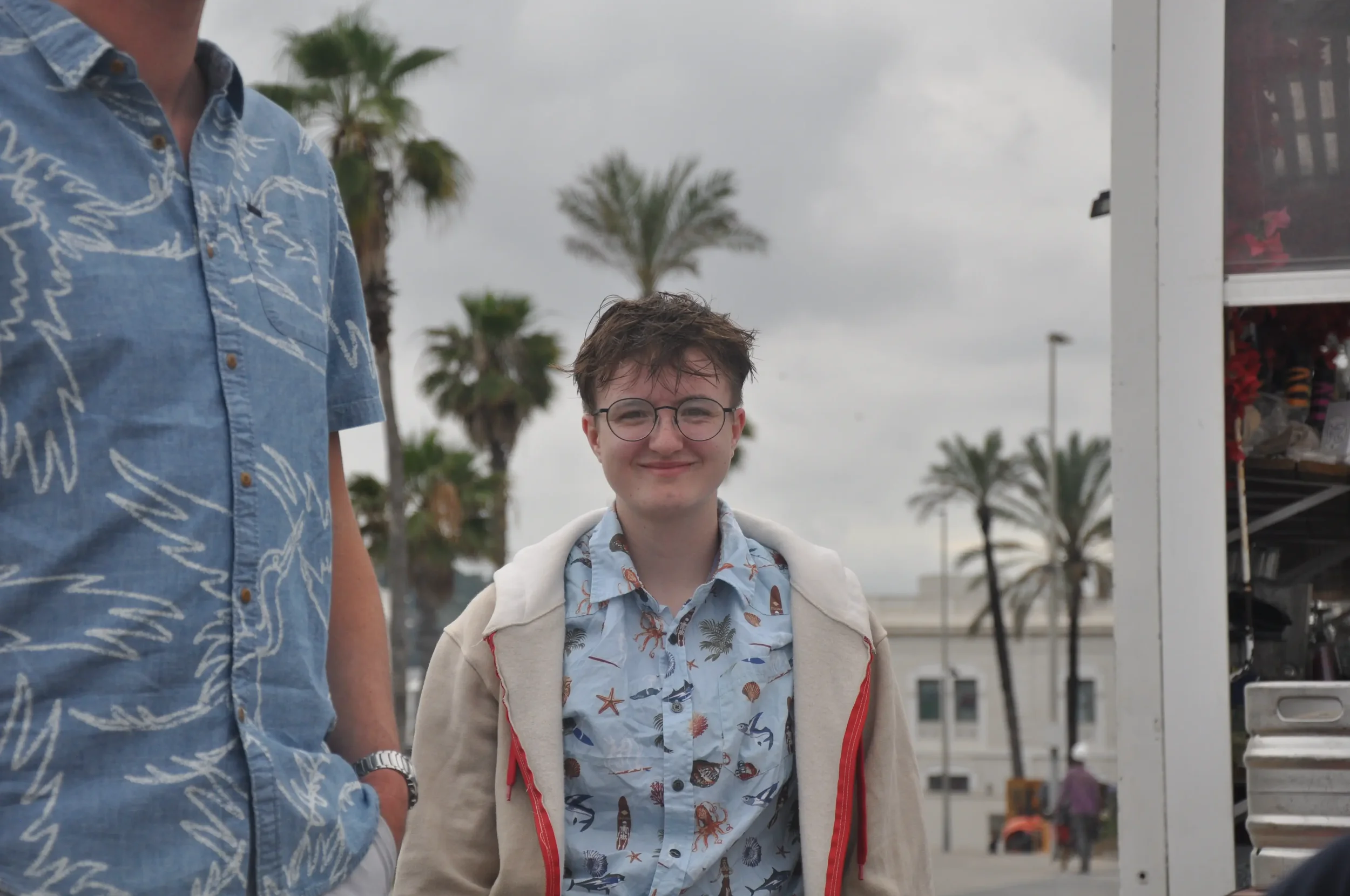 A young man smiling at camera, wearing glasses, a light blue shirt with sea creatures, and a beige jacket with a hood, standing outdoors near palm trees and a food stand on gray, overcast day.
