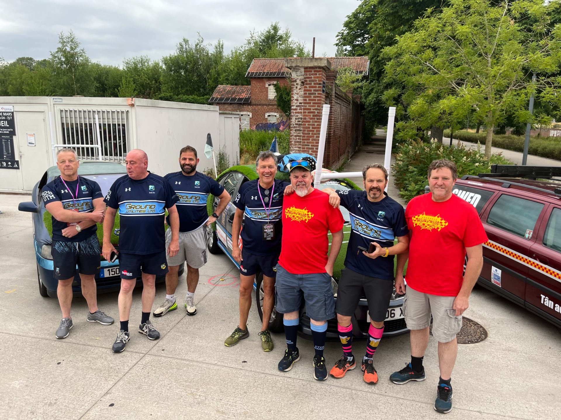 Group of seven men standing outdoors, some wearing matching blue and black shirts, two in red shirts, smiling for the photo, with cars and greenery in the background.