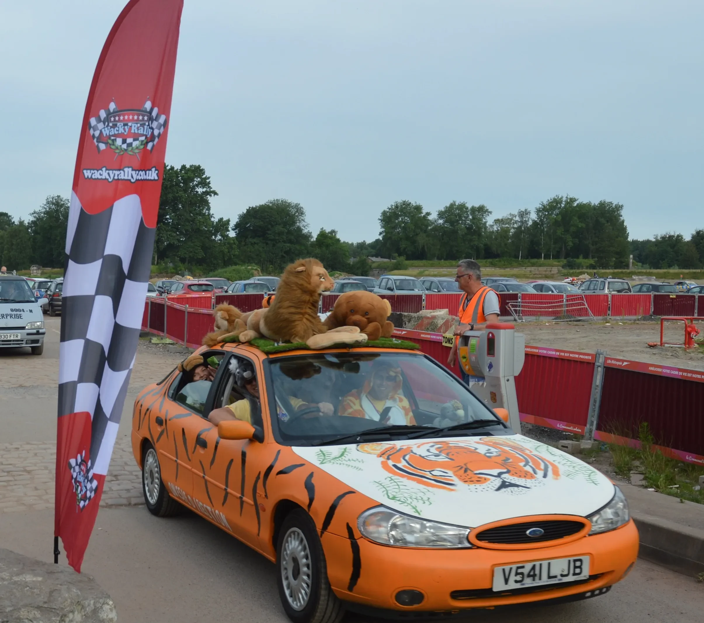 Orange car decorated with tiger stripes and a tiger face on the hood, with stuffed animals of a lion, teddy bear, and a dog on top, at a rally event.