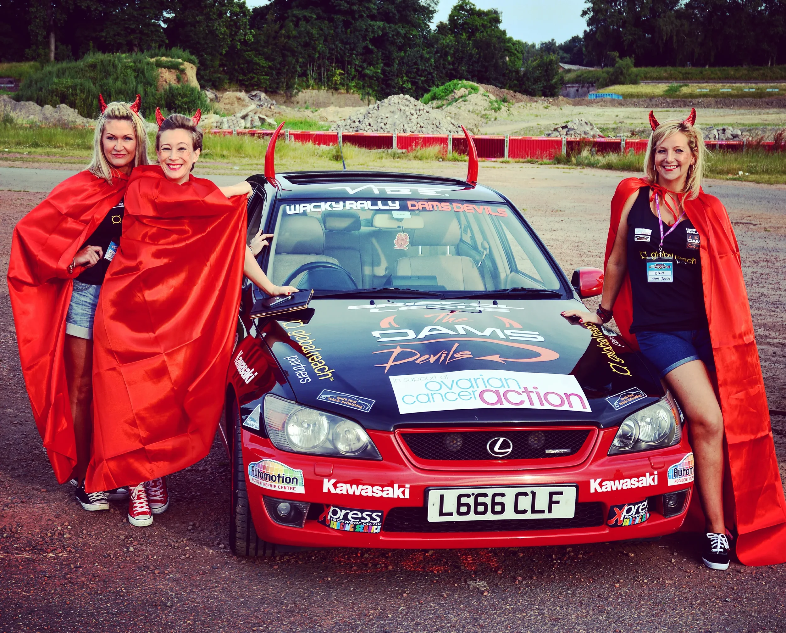 Three women dressed as devils with red horns, red capes, and black shirts pose around a rally car decorated for a charity event supporting ovarian cancer awareness, on an outdoor dirt lot with greenery in the background.