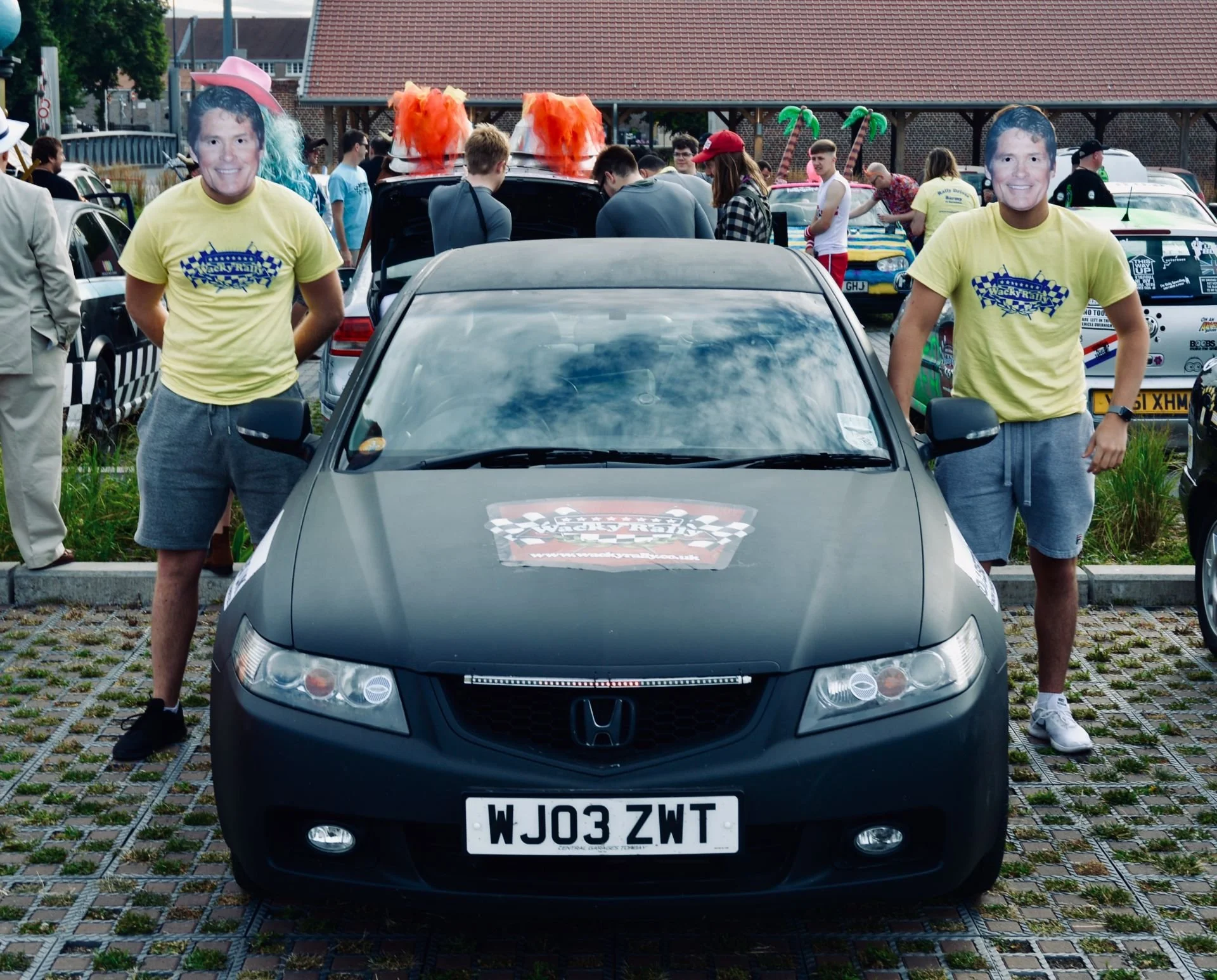 Black Honda car with a rally sticker on the hood, surrounded by people attending a car event, with two men in yellow rally shirts standing next to it.