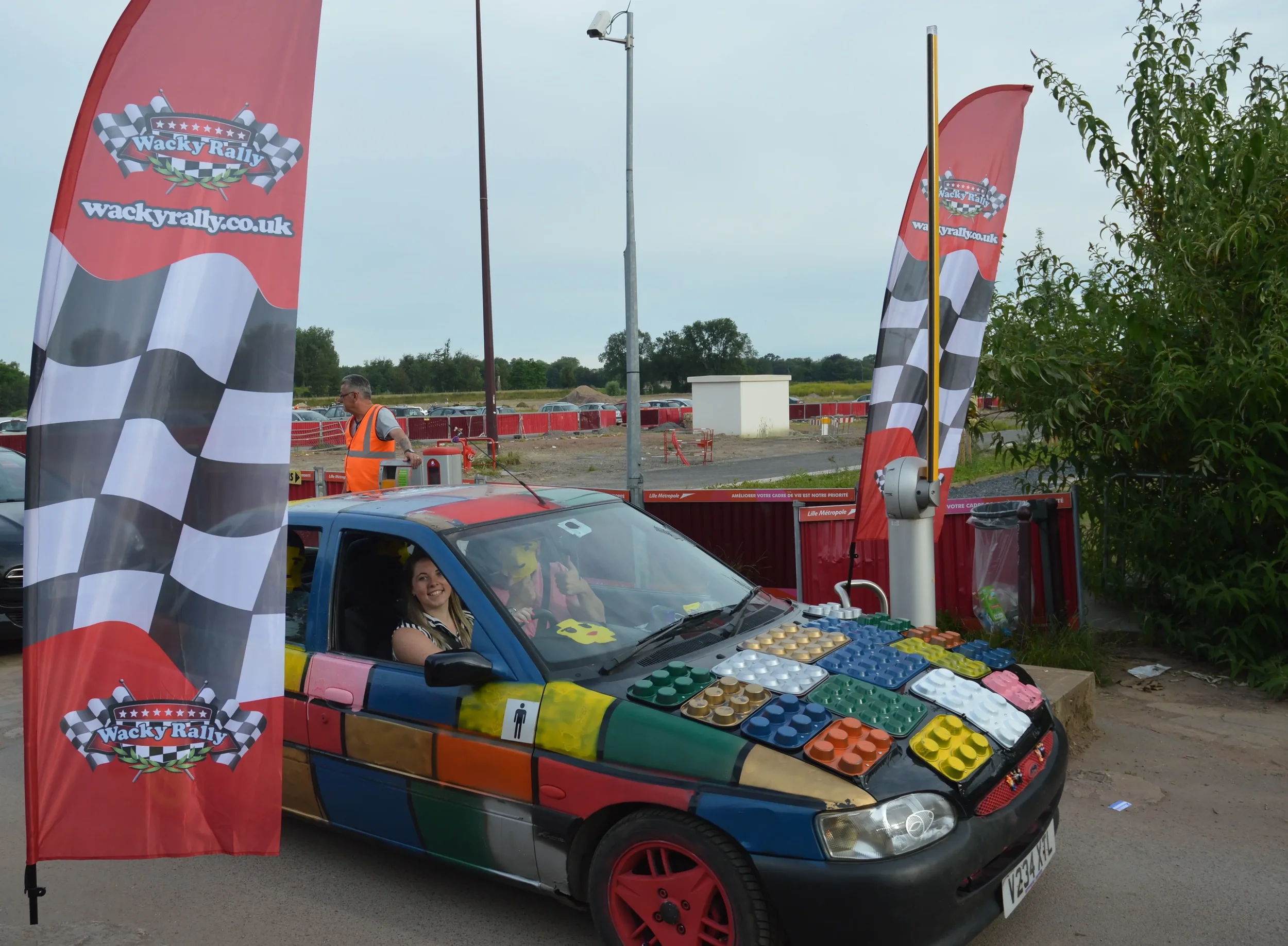A colorful car decorated with various LEGO pieces on the hood and sides, parked at a rally event with Wacky Rally flags nearby and people in the car, smiling and taking photos.