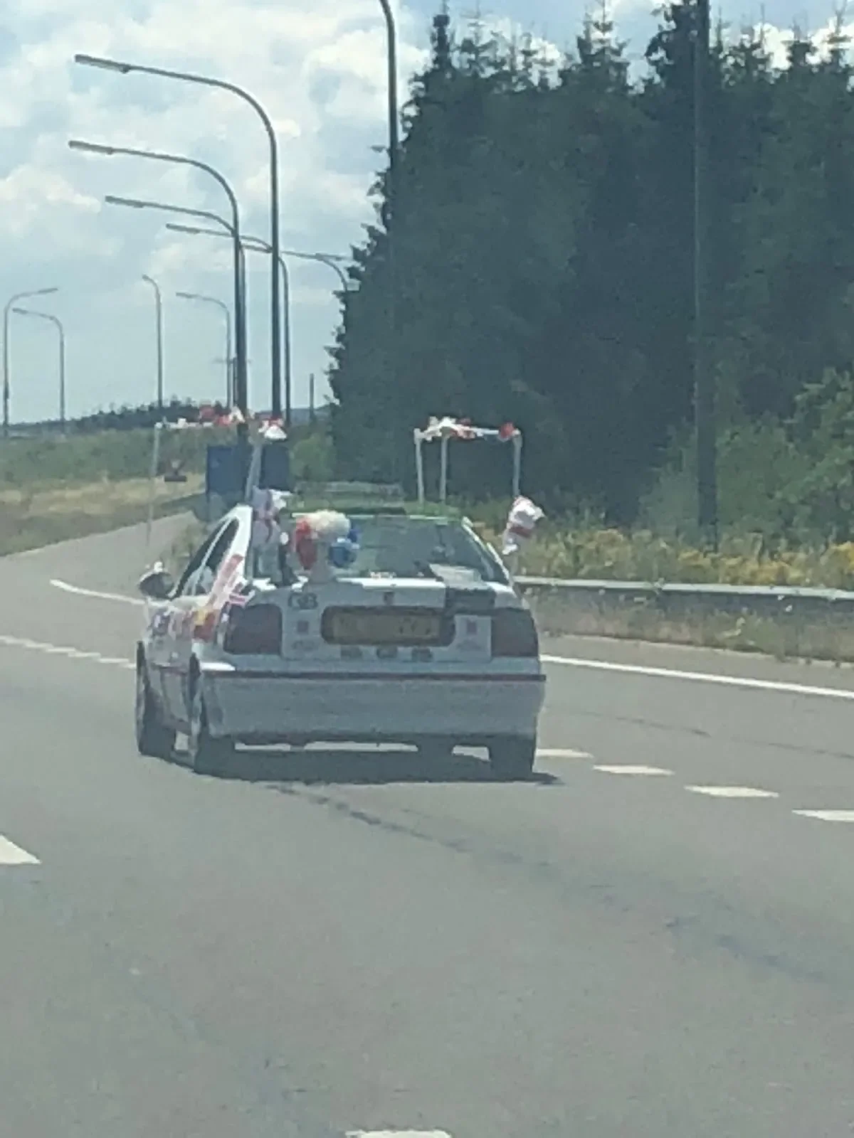 A car decorated with American flags and patriotic items, driving on a highway with streetlights and trees in the background.