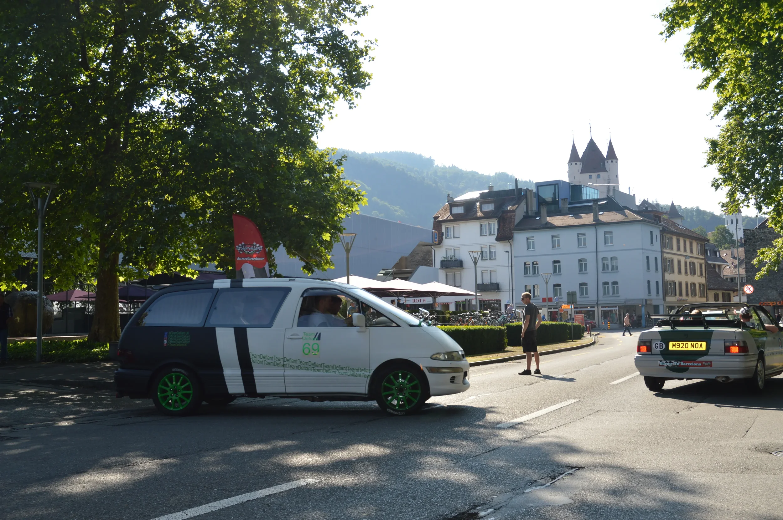 An urban street scene with a white and black car with green wheels in the foreground, people walking on the sidewalk, and a castle-like building on a hill in the background, surrounded by trees and mountains.