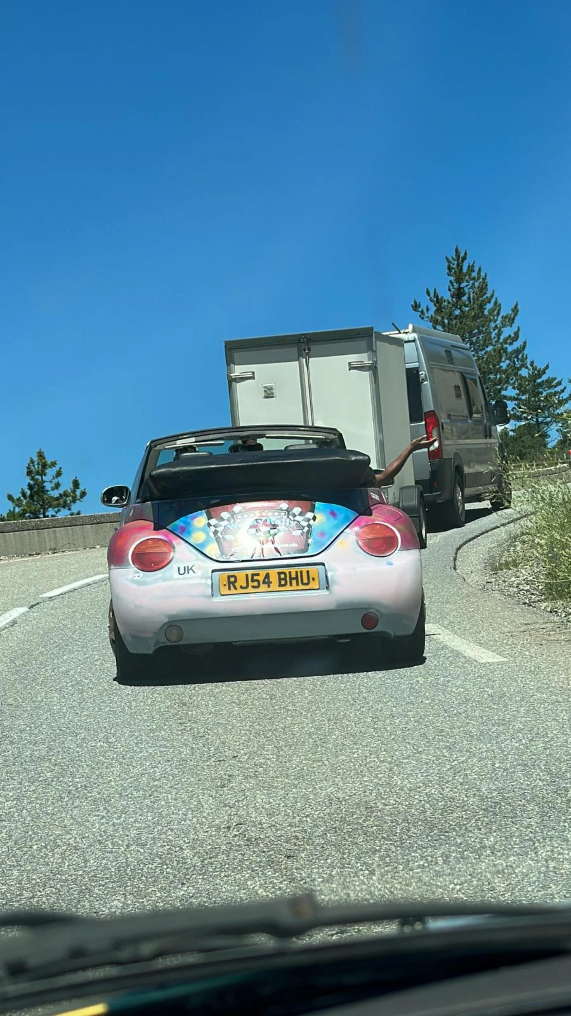 A small convertible car with a colorful graphic on the back, following a delivery van on a mountain road under a clear blue sky.