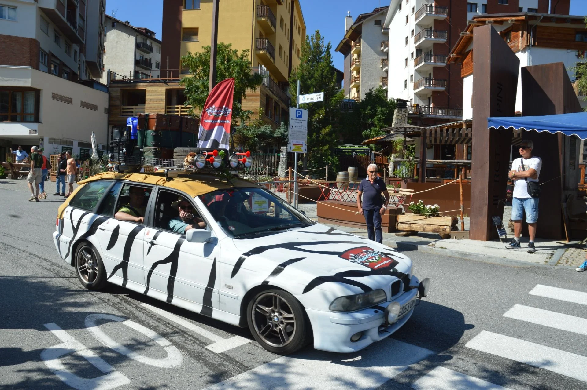 A white car with black zebra-like stripes and a lion crest on the hood is driving through a city street. People stand on the sidewalk, and tall buildings are visible in the background.