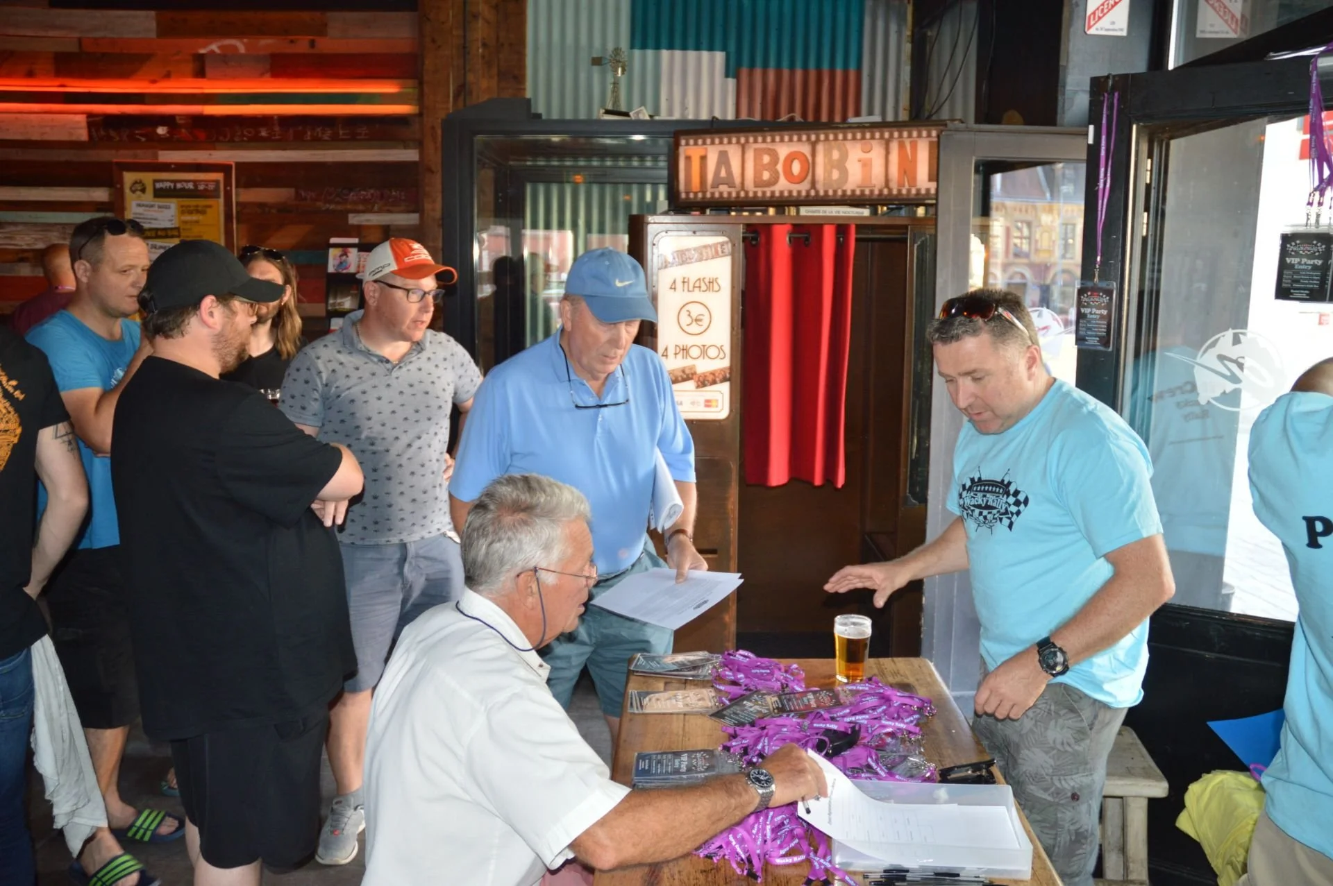 People gathered around a table at an event, receiving medals and paperwork, outside a venue with a sign that reads 'TABOBIN' and promotional posters on the wall.