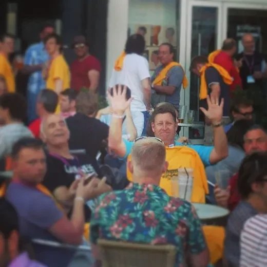 A group of people gathered outdoors, some sitting at tables and others standing, with a man in a blue shirt and yellow scarf waving and smiling at the camera.