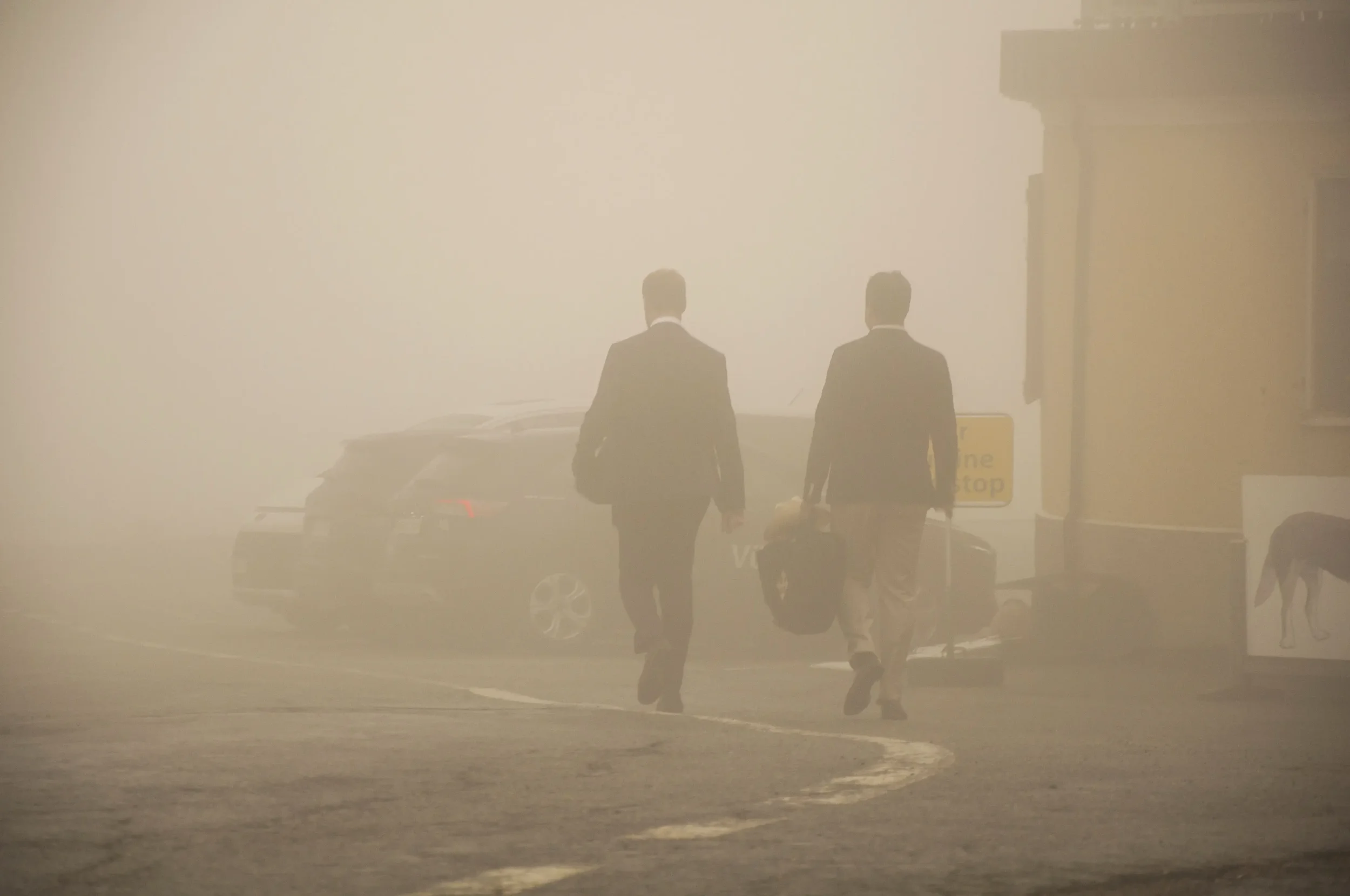 Two men in business suits walking through a thick fog on a city street, one carrying a briefcase and the other a bag.