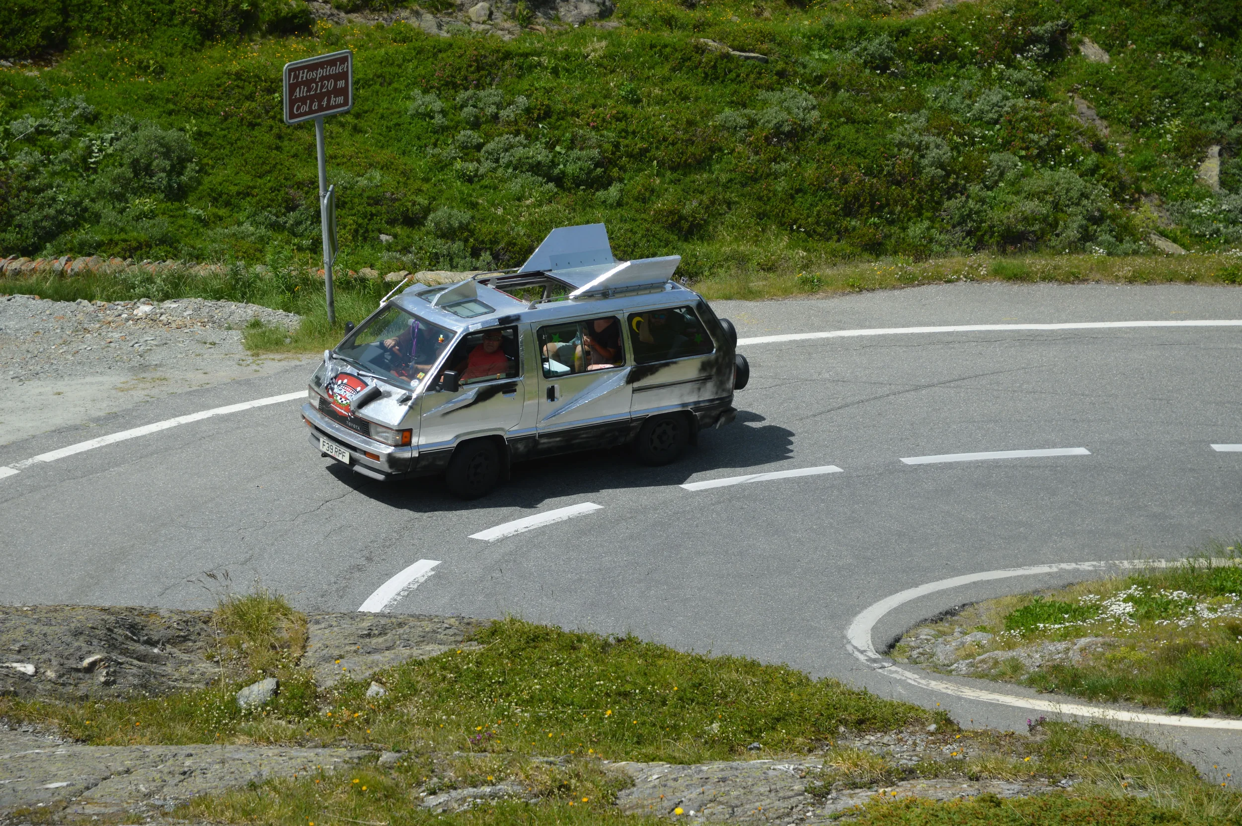 A modified silver van with black stripes on a mountain road, with a sign indicating the altitude and distance to a hospital, surrounded by greenery and rocks.