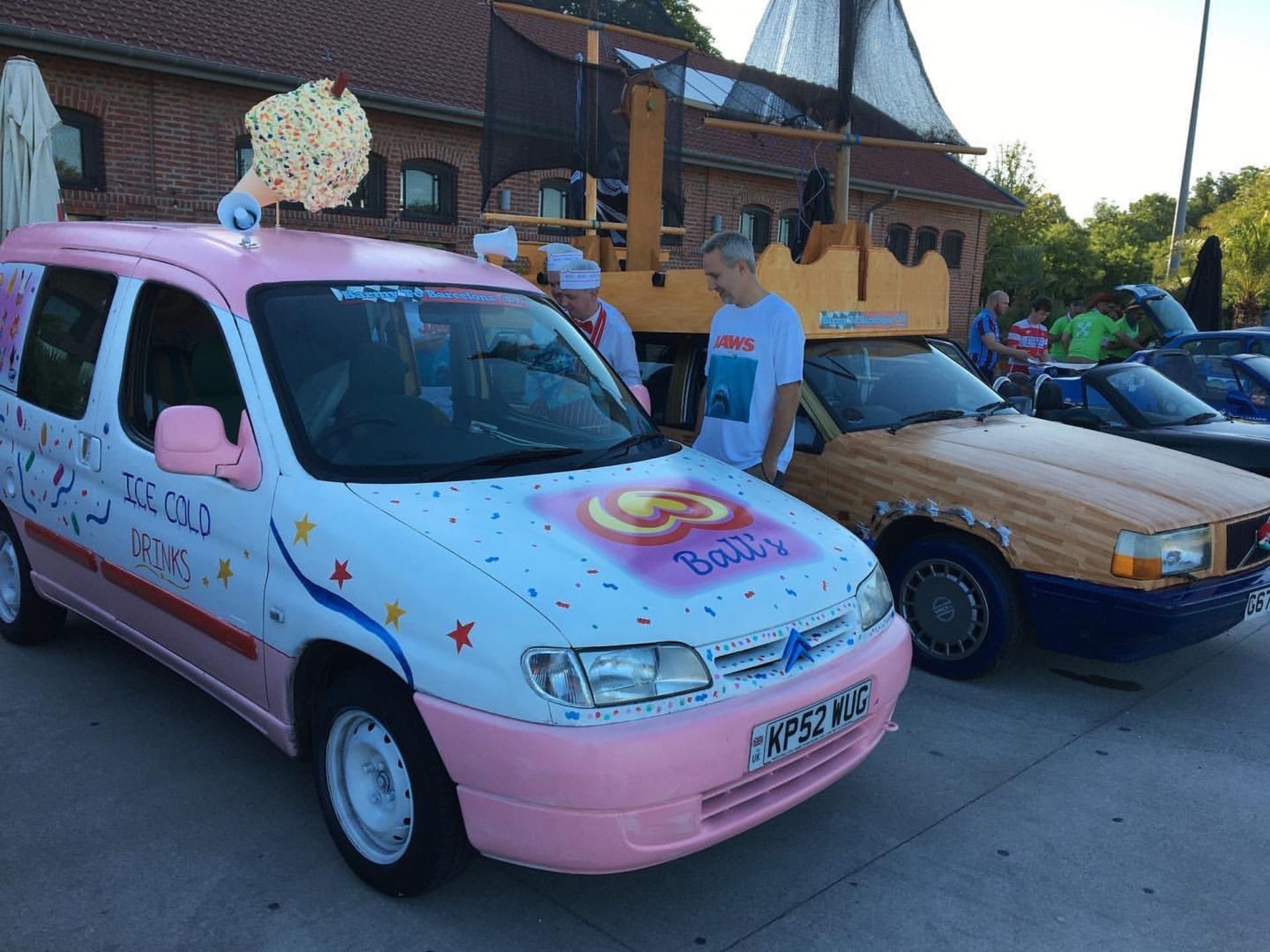 A pink ice cream truck decorated with multicolored sprinkles and stars, labeled 'Ice Cold Drinks' and 'Baby', parked next to a wooden Cars-themed car at an outdoor event. People are standing nearby.