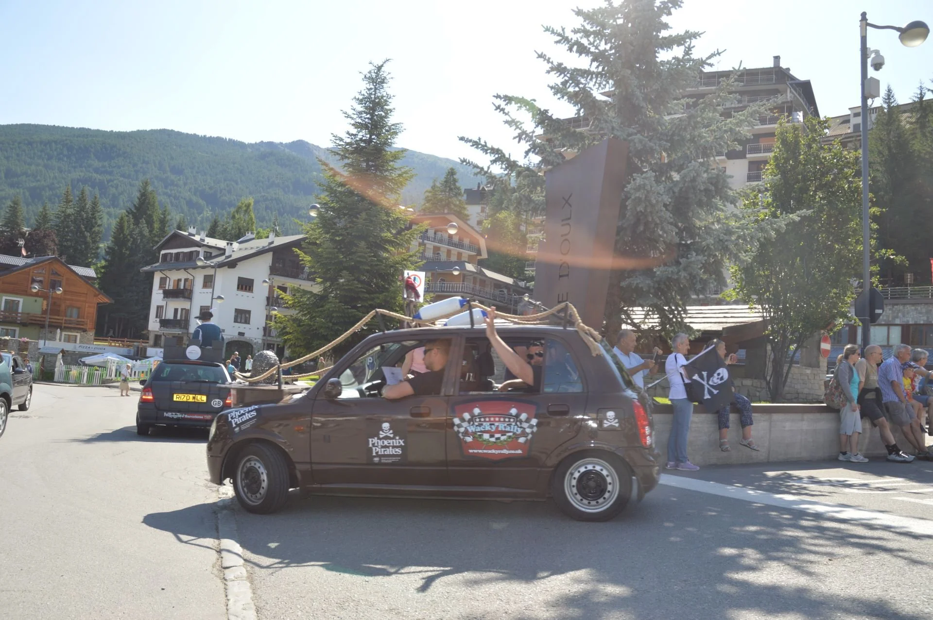 A small brown car with 'Phoenix Pirates' and 'Wacky Rally' decals driven by two men dressed as pirates, one waving from the window, as part of a parade with people lining the street, in a mountainous town with chalet-style buildings and trees.