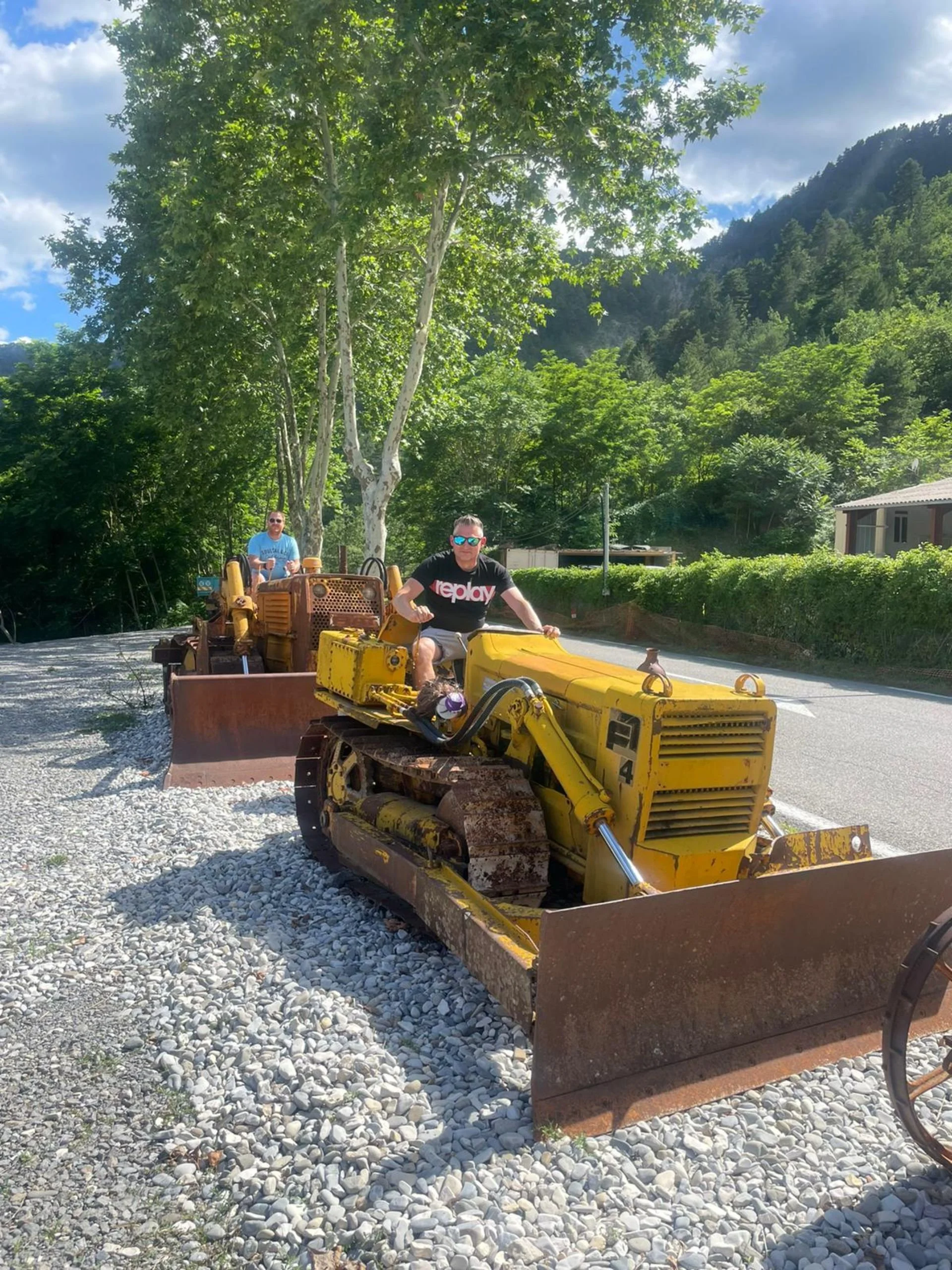 Two people operating vintage bulldozers on a gravel surface surrounded by green trees and mountainous landscape, under a partly cloudy sky.