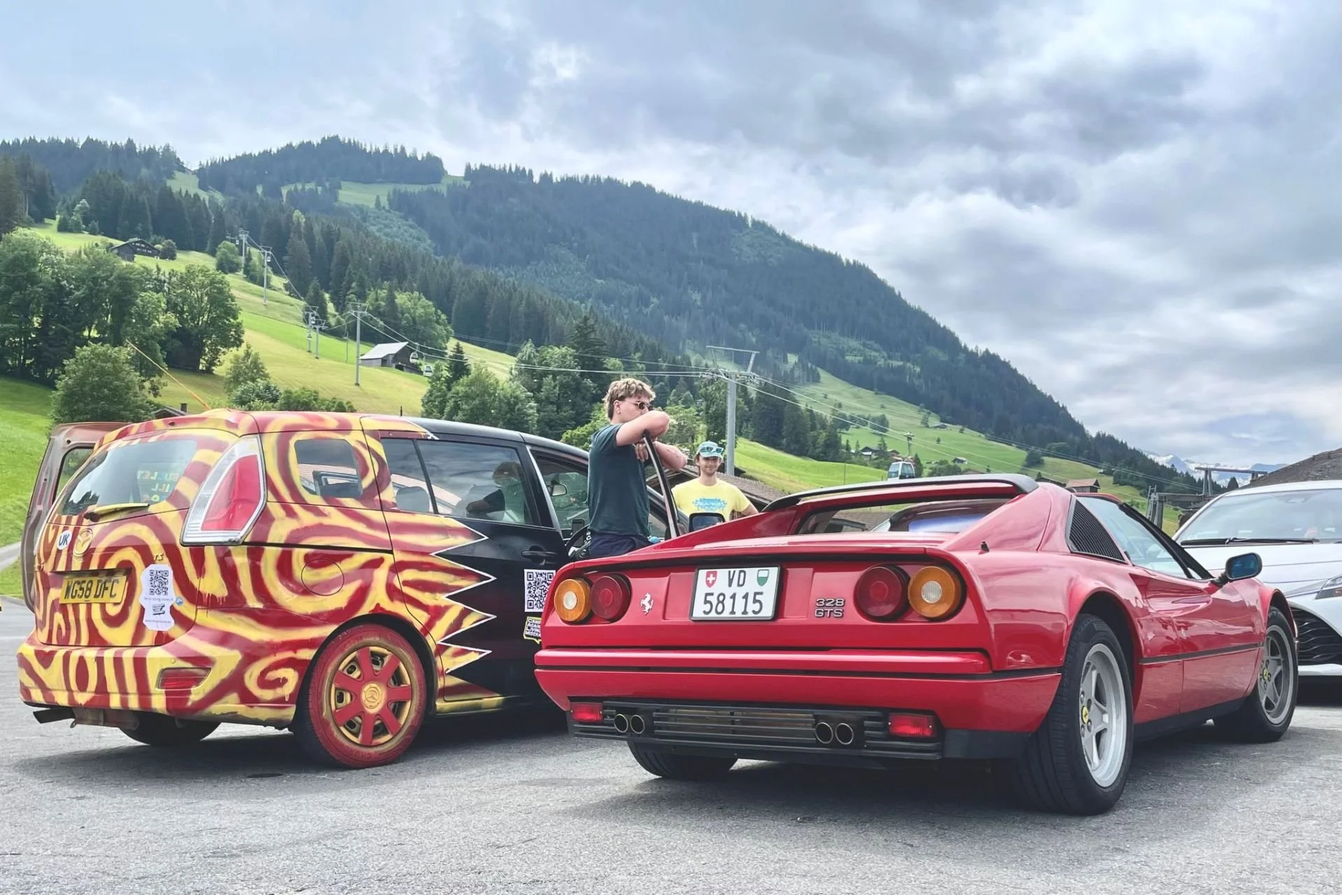 Two cars, one with a bright, funky painted pattern and the other a classic red Ferrari, parked outdoors with green hills and mountains in the background, and two young men standing between the cars, one adjusting his sunglasses.