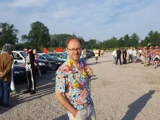 A man wearing glasses, a colorful floral shirt, and a lei, standing outdoors at a crowded event with people, cars, and tents in the background.