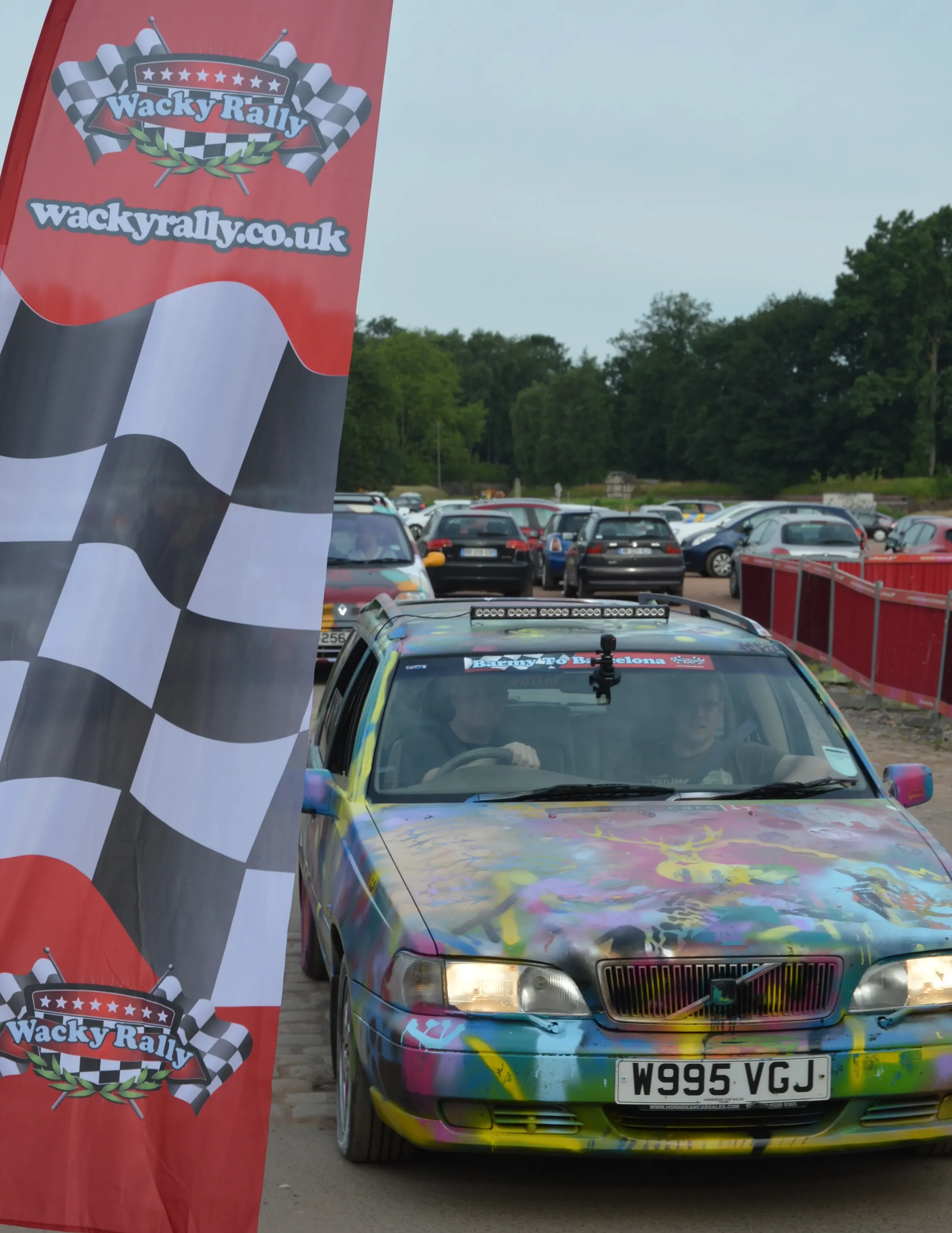 A colorful, graffiti-style painted car at a rally event, parked beside a Wacky Rally banner with a checkered flag design.