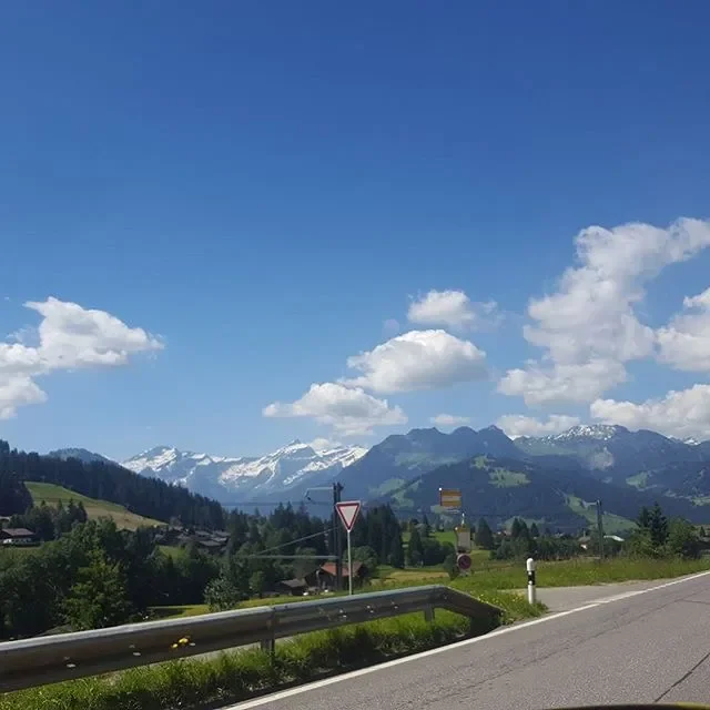 Scenic view of snow-capped mountains under blue sky with fluffy clouds, surrounded by green hills and trees, taken from a roadside.