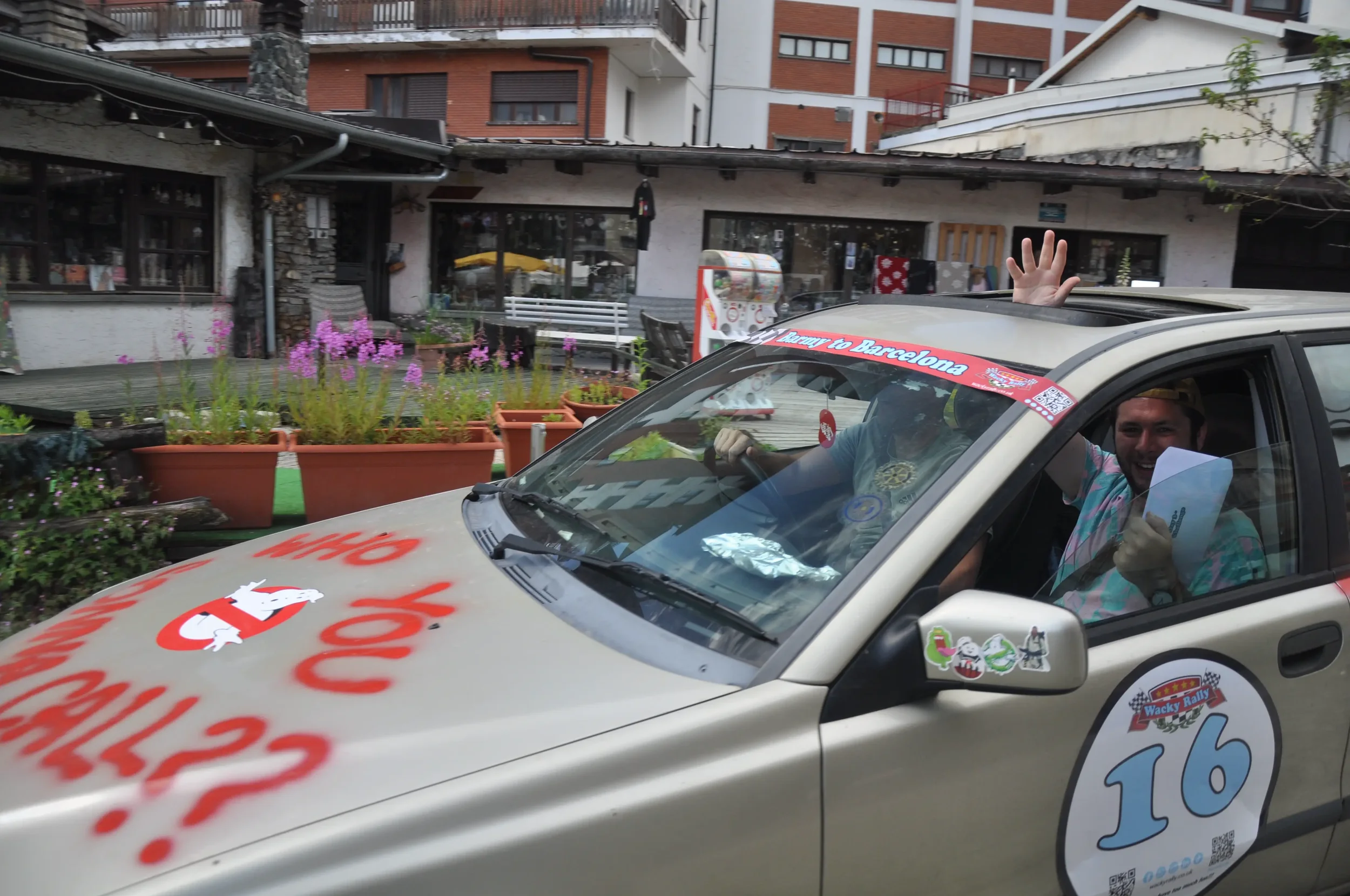 Two men inside a rally car, one waving through the sunroof and smiling, the other giving a thumbs-up. The car is decorated with stickers, including a large number 16 and racing-themed logos, parked in front of a building with plants and pink flowers.