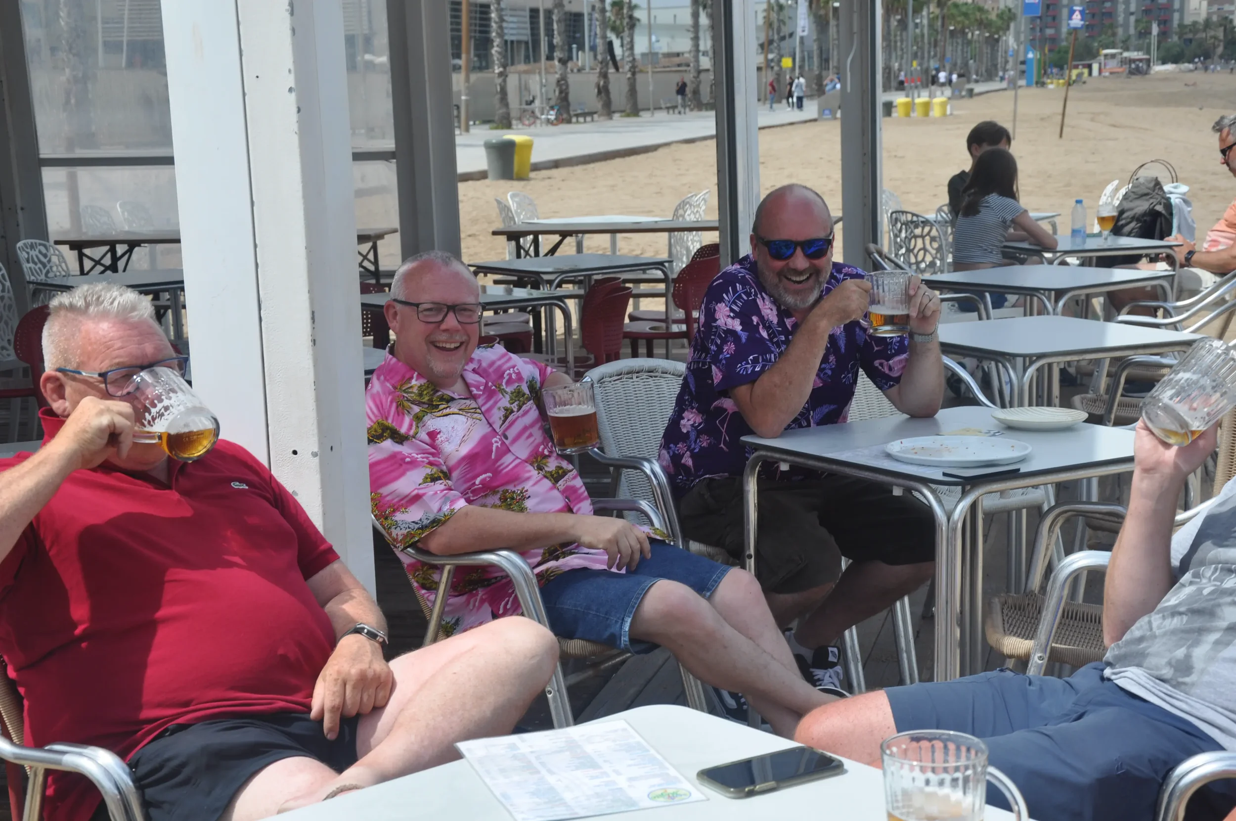 Four men sitting at outdoor bar or restaurant, enjoying drinks and smiling, with a sandy beach andboardwalk in the background.