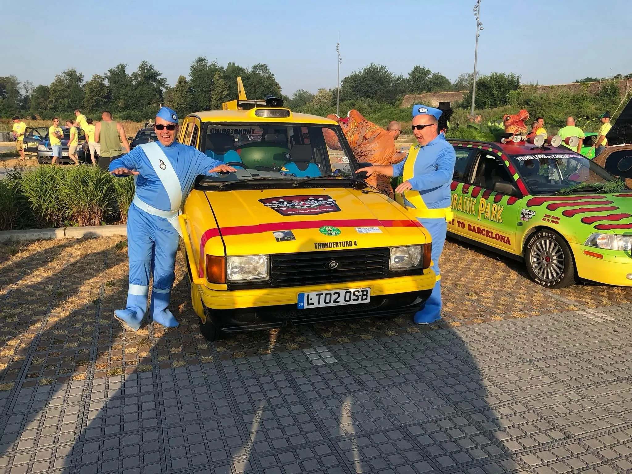 Two people dressed as paramedics standing next to a yellow car with racing decals at a car show. There is another decorated car behind them and a crowd of people in the background.