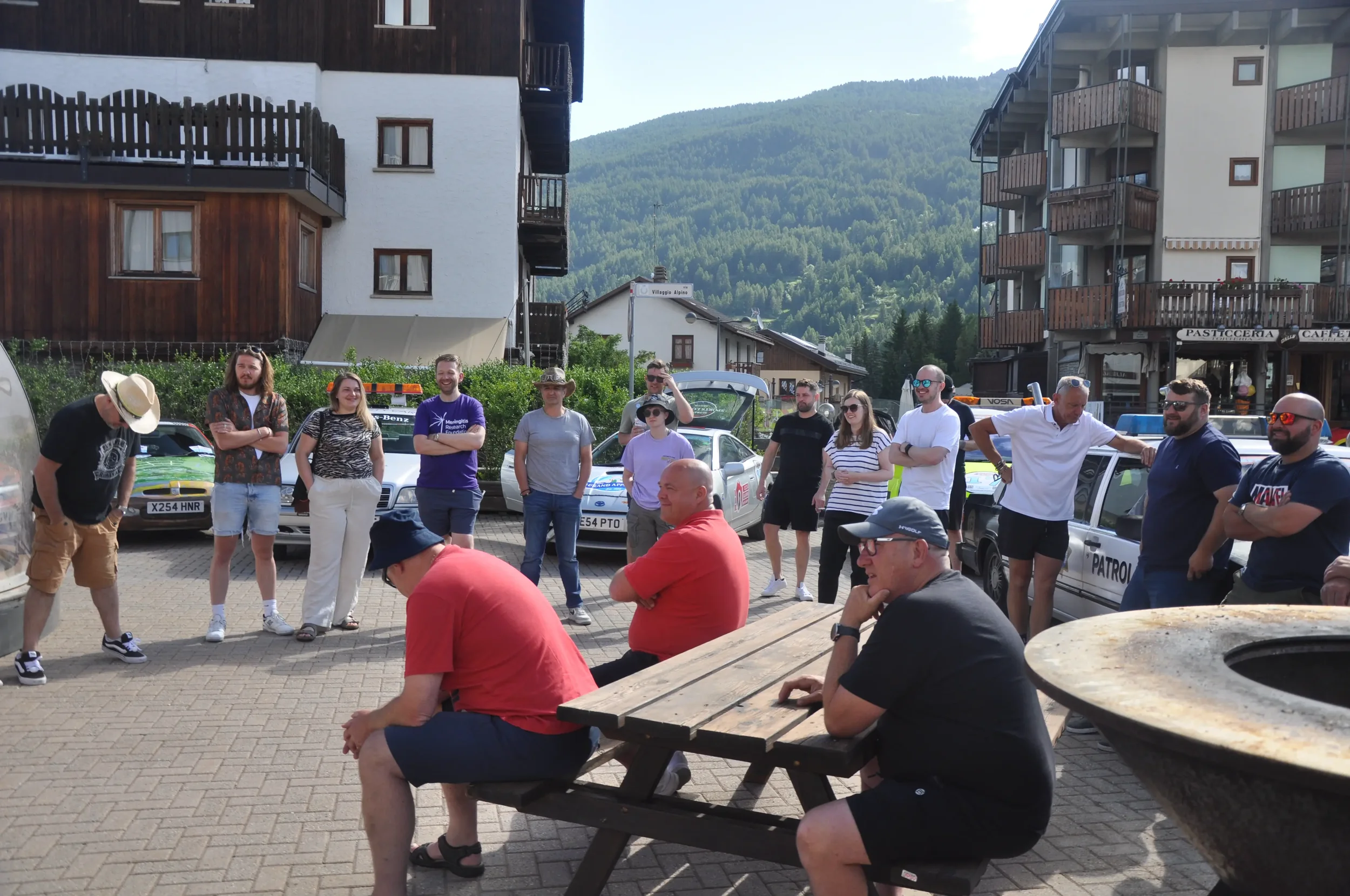 Group of people gathered outdoors in a mountain town, some sitting at a picnic table and others standing, during sunny weather.